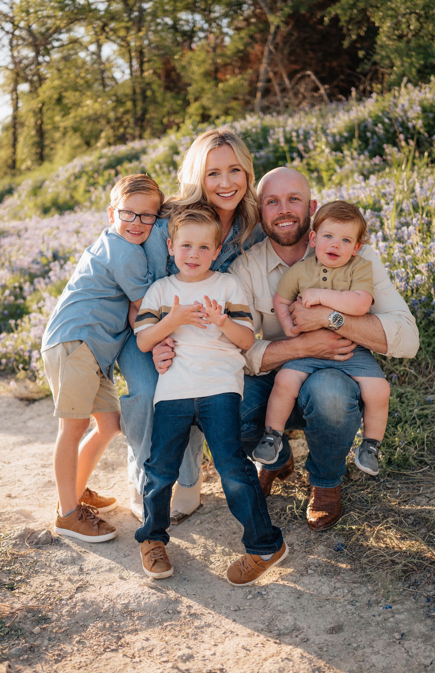 Close family portrait with group hugging and smiling amidst blooming wildflowers on a sunny hillside.