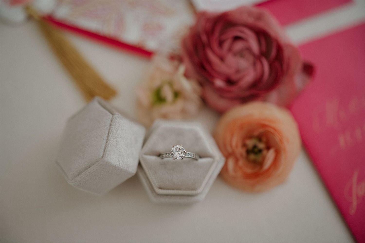 A sparkling diamond engagement ring sits in a white hexagonal box surrounded by soft pink and peach ranunculus flowers.