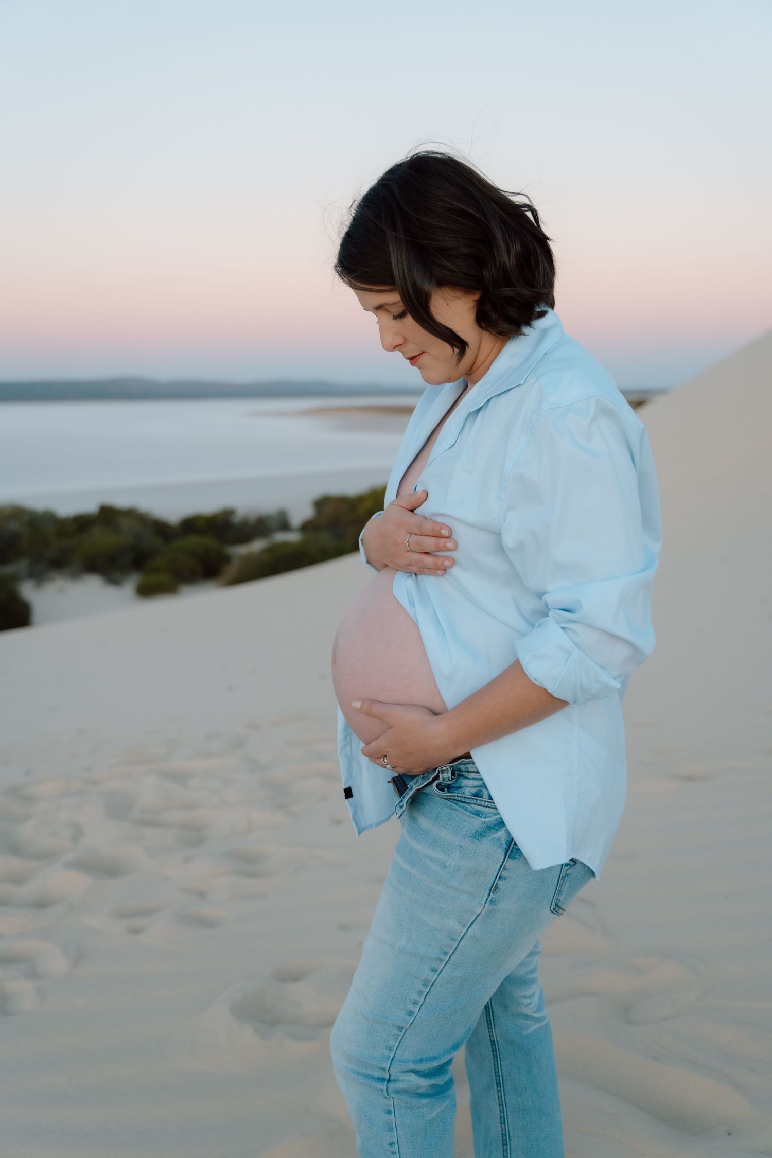 Pregnancy photos captured on beach with person in chambray shirt and jeans against sunset sky.