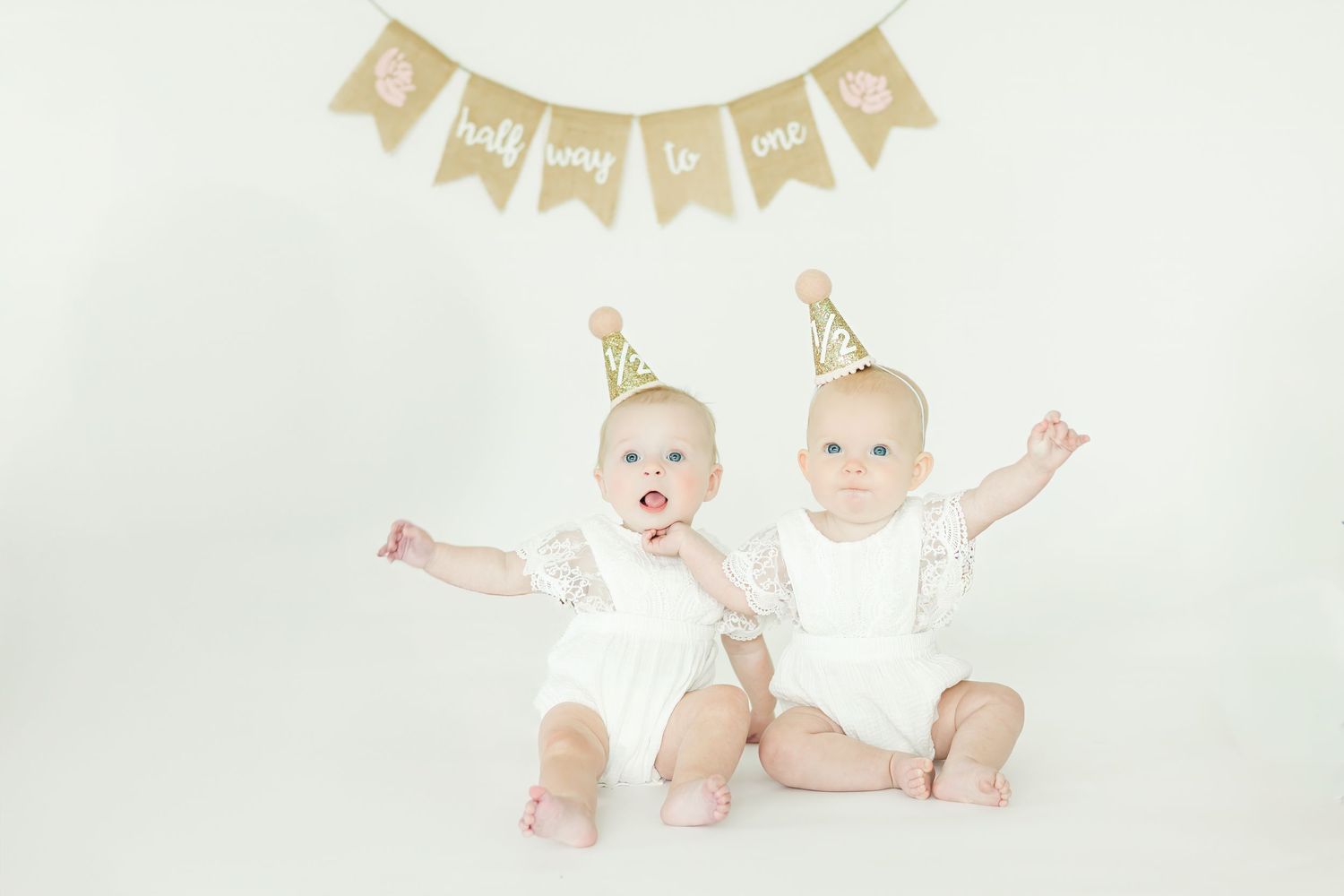 Twin babies in white outfits lying under decorative banner during milestone photo session.