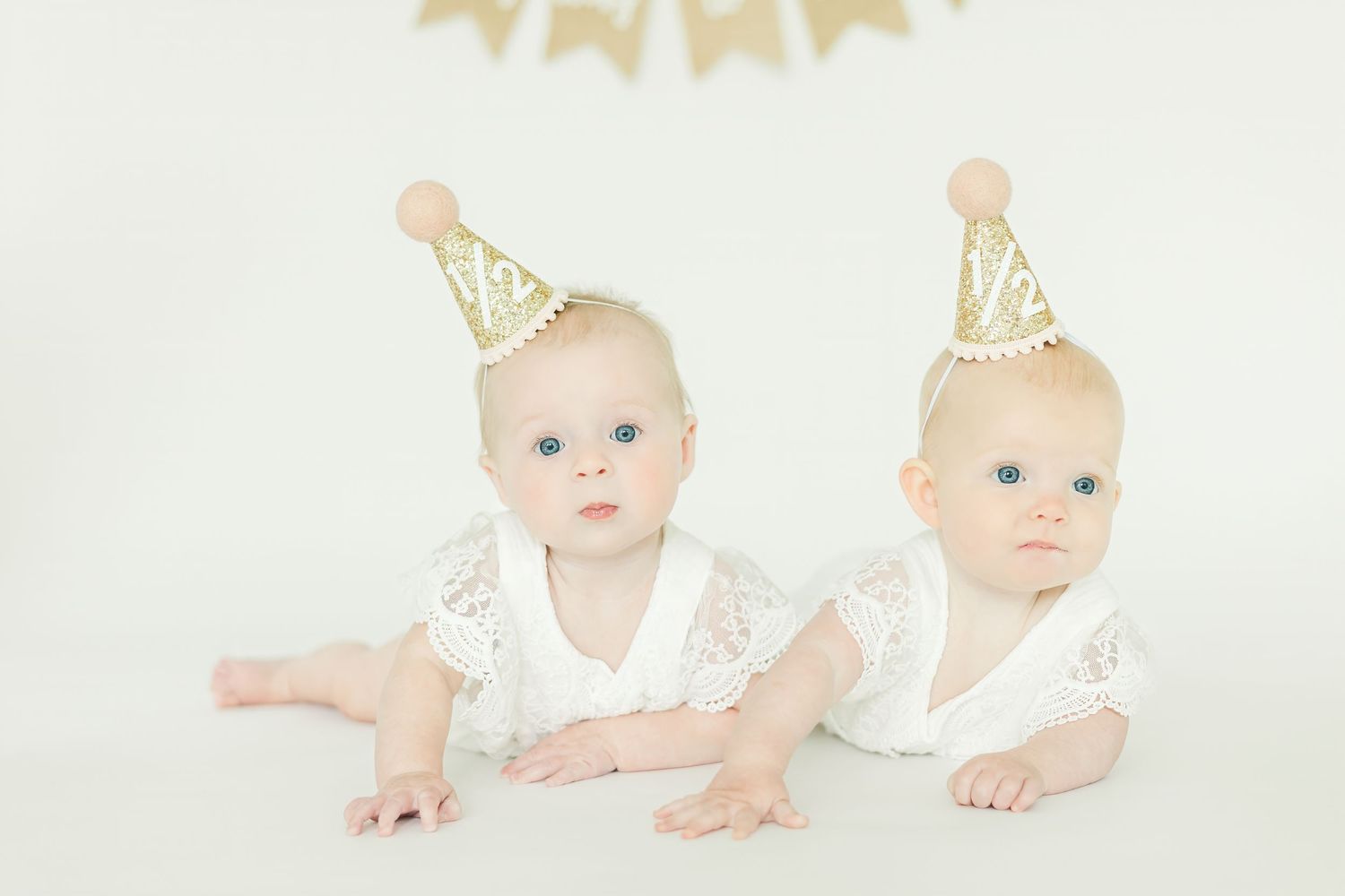 Twin babies in matching white outfits with gold party hats crawling on white backdrop.