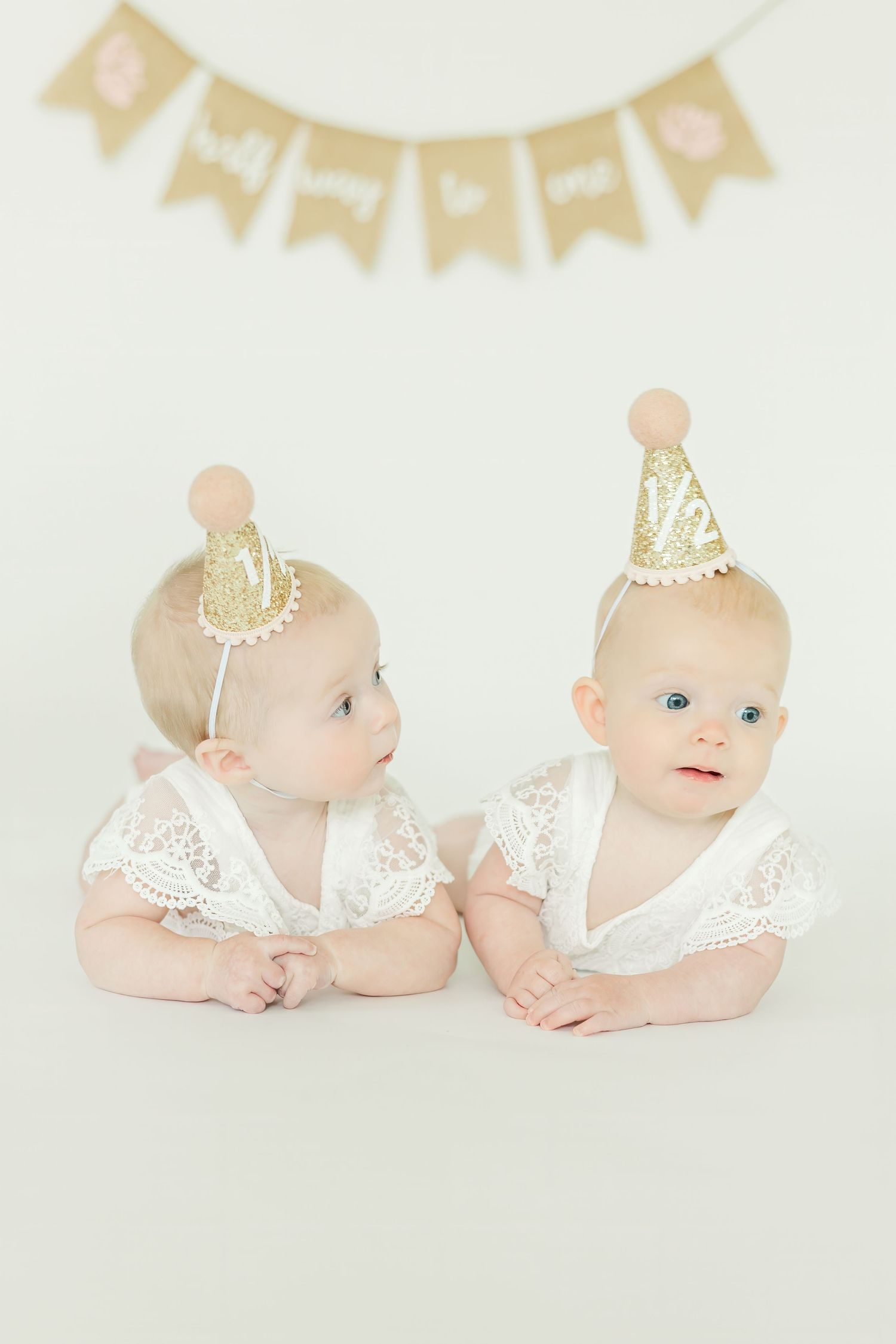 Baby wearing white lace outfit and gold crown lying on white background with banner decorations.