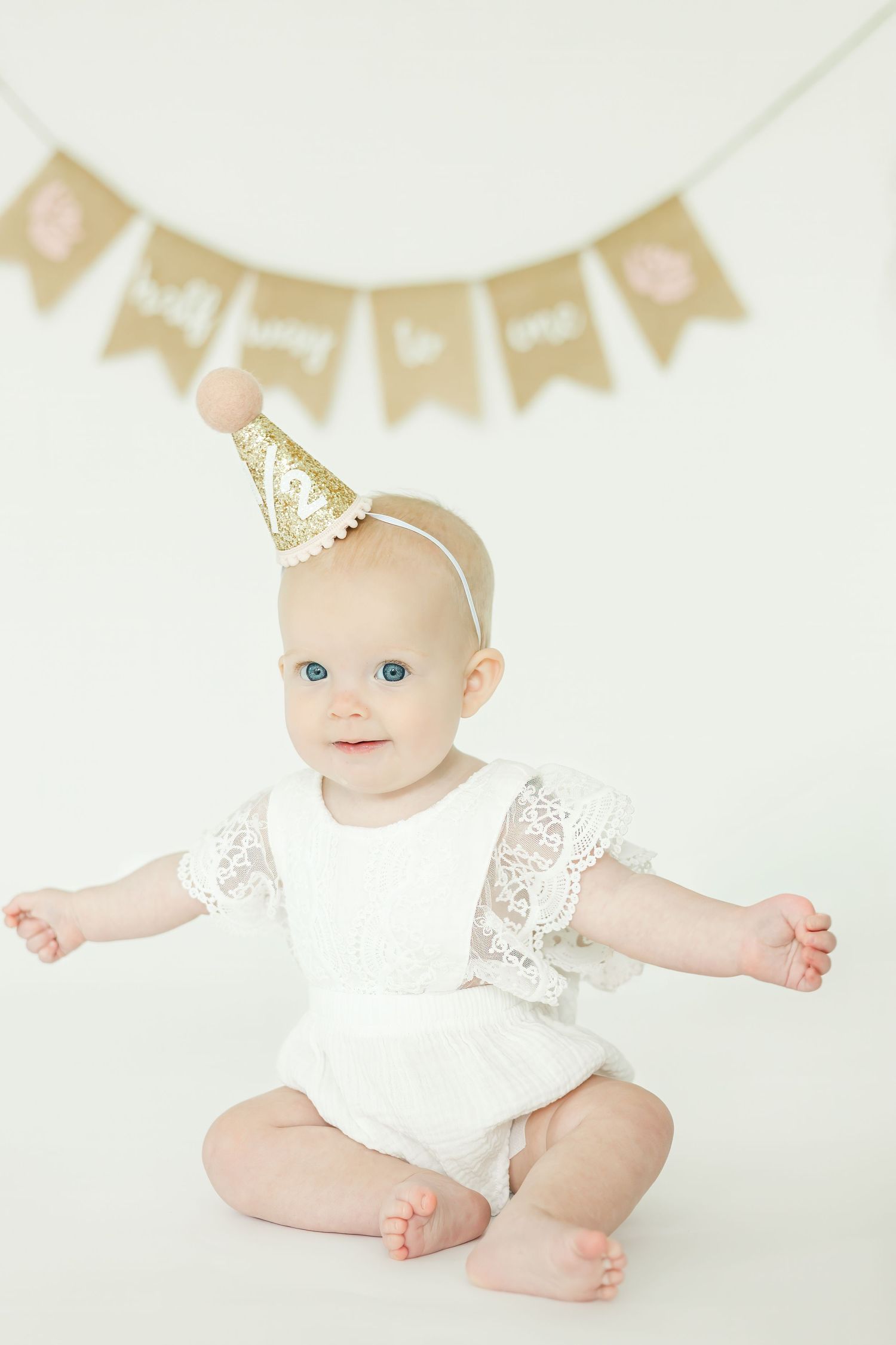 Baby in white lace dress and gold crown posing on light backdrop.