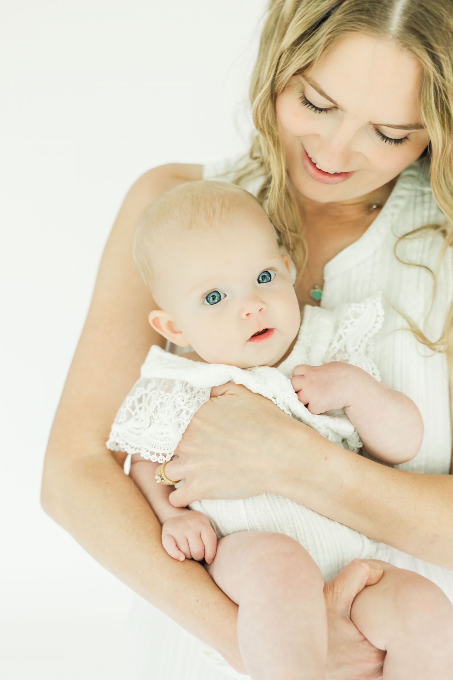 Tender moment between mother and baby in white lace outfits against light background.