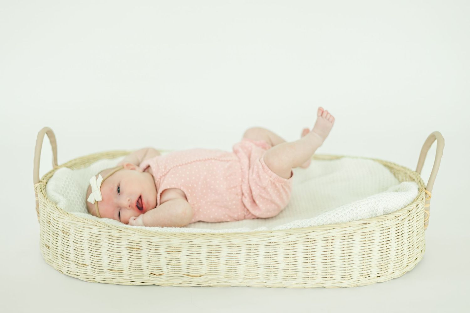 Infant wearing soft pink onesie lying in white wicker bassinet during photo session.