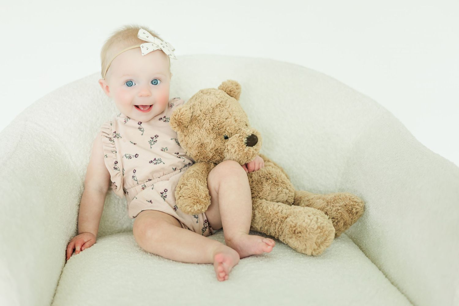Baby in floral outfit cuddling with teddy bear on white couch.