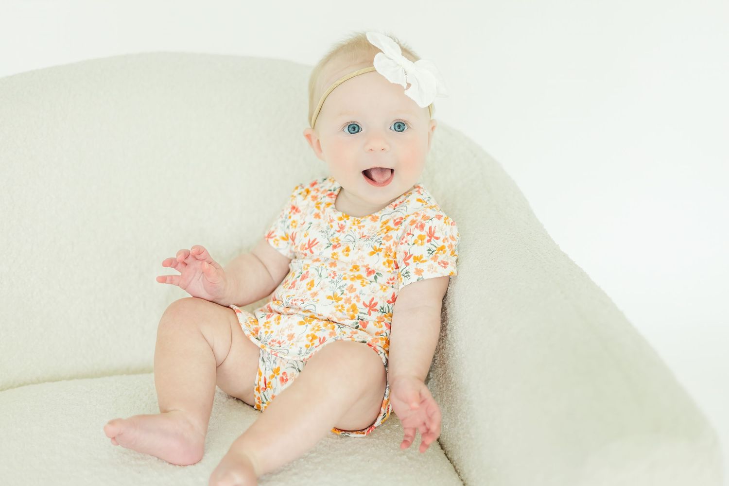 Baby in floral romper smiling and playing on light colored sofa.