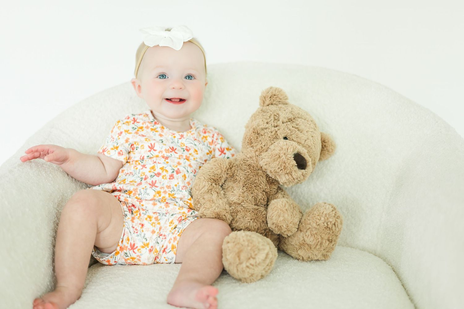 Baby in floral outfit rests on white surface next to teddy bear.