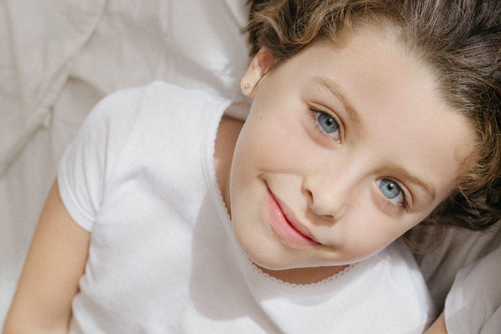 Primer plano retrato de un niña con una camiseta blanca acostado sobre ropa de cama de color claro.