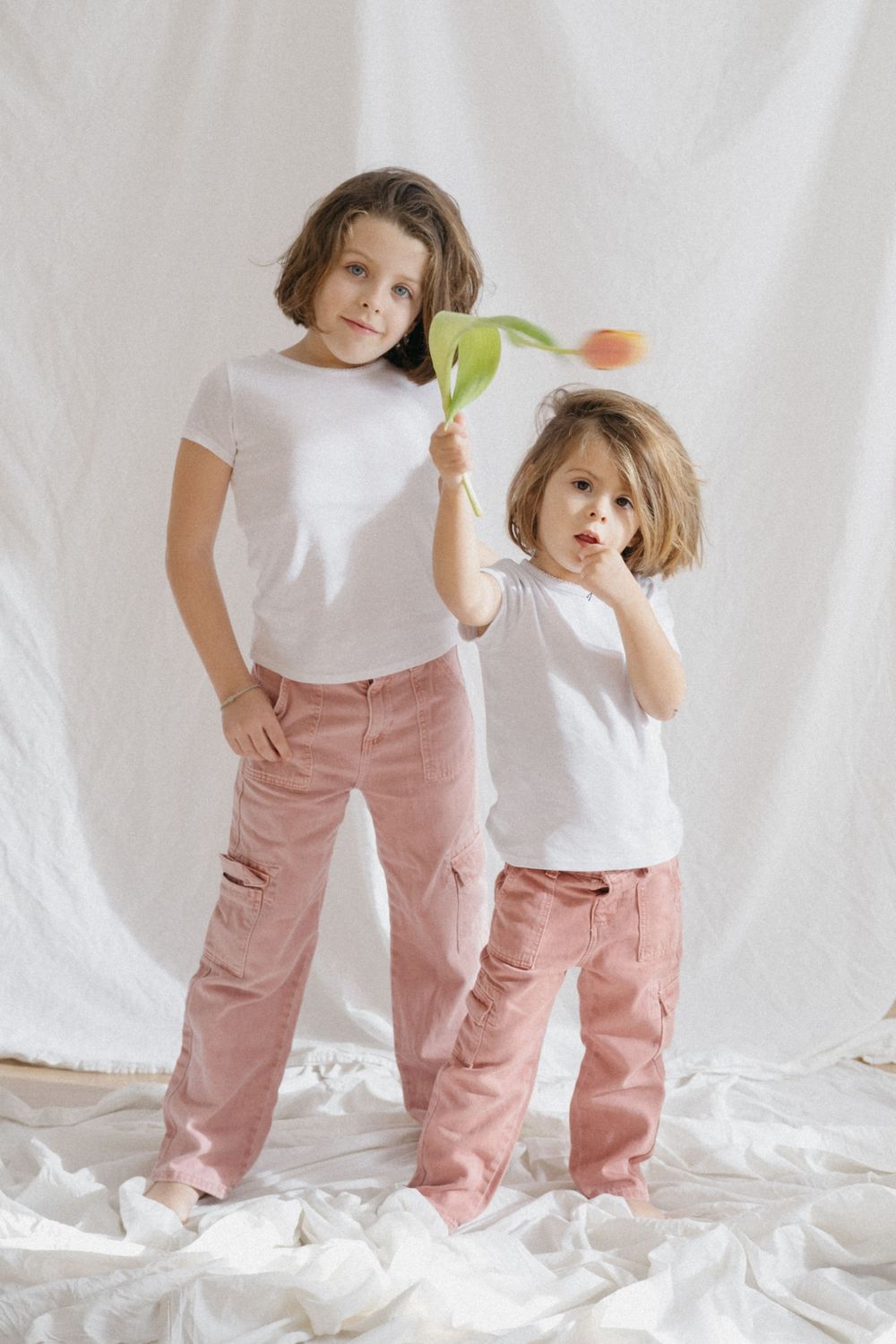 Dos niñas con camisas blancas y pantalones rosas jugando con flor sobre fondo blanco.
