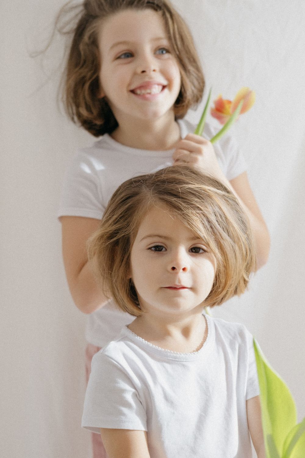 Dos niños con camisetas blancas sonriendo sobre un fondo claro con una flor de primavera.