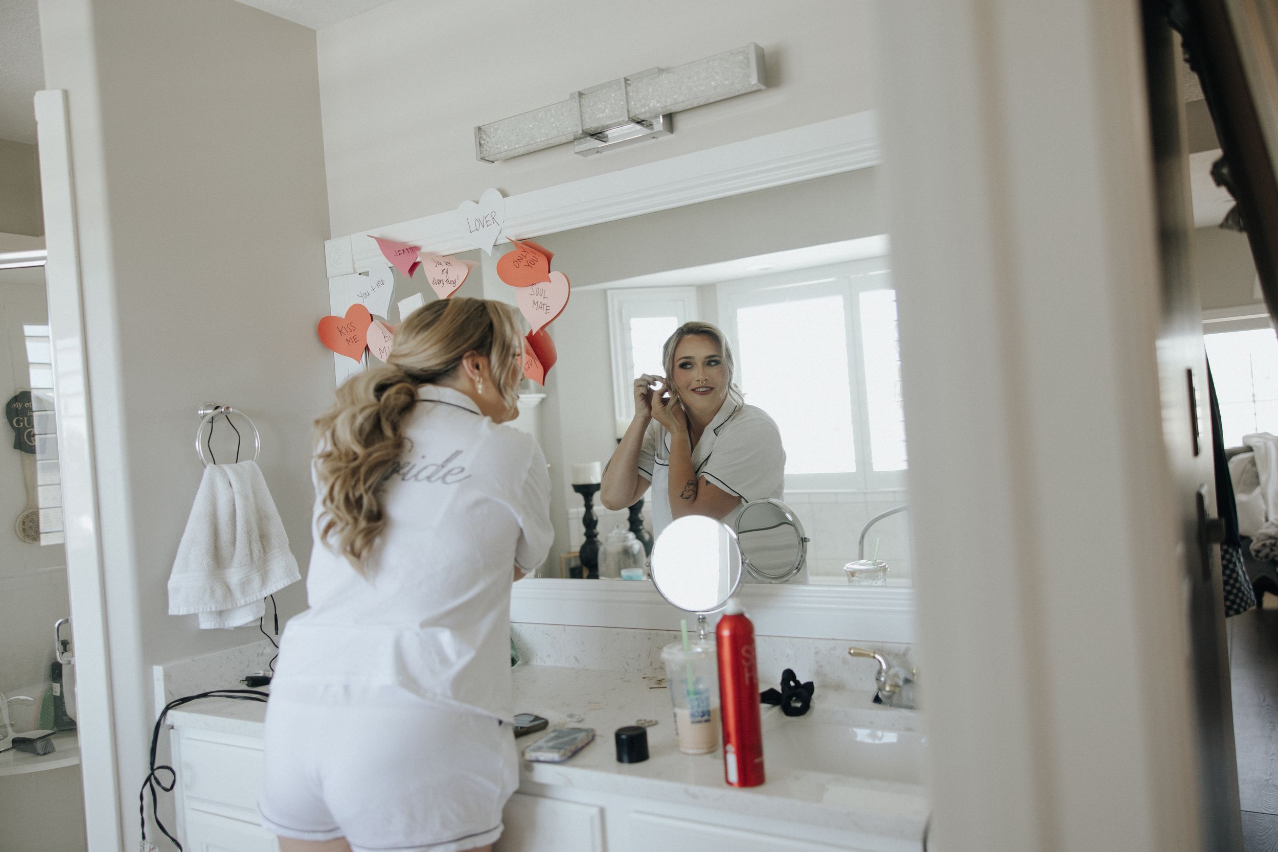 Figure in white robe applies makeup at bathroom vanity with mirrors and natural light streaming in through window.