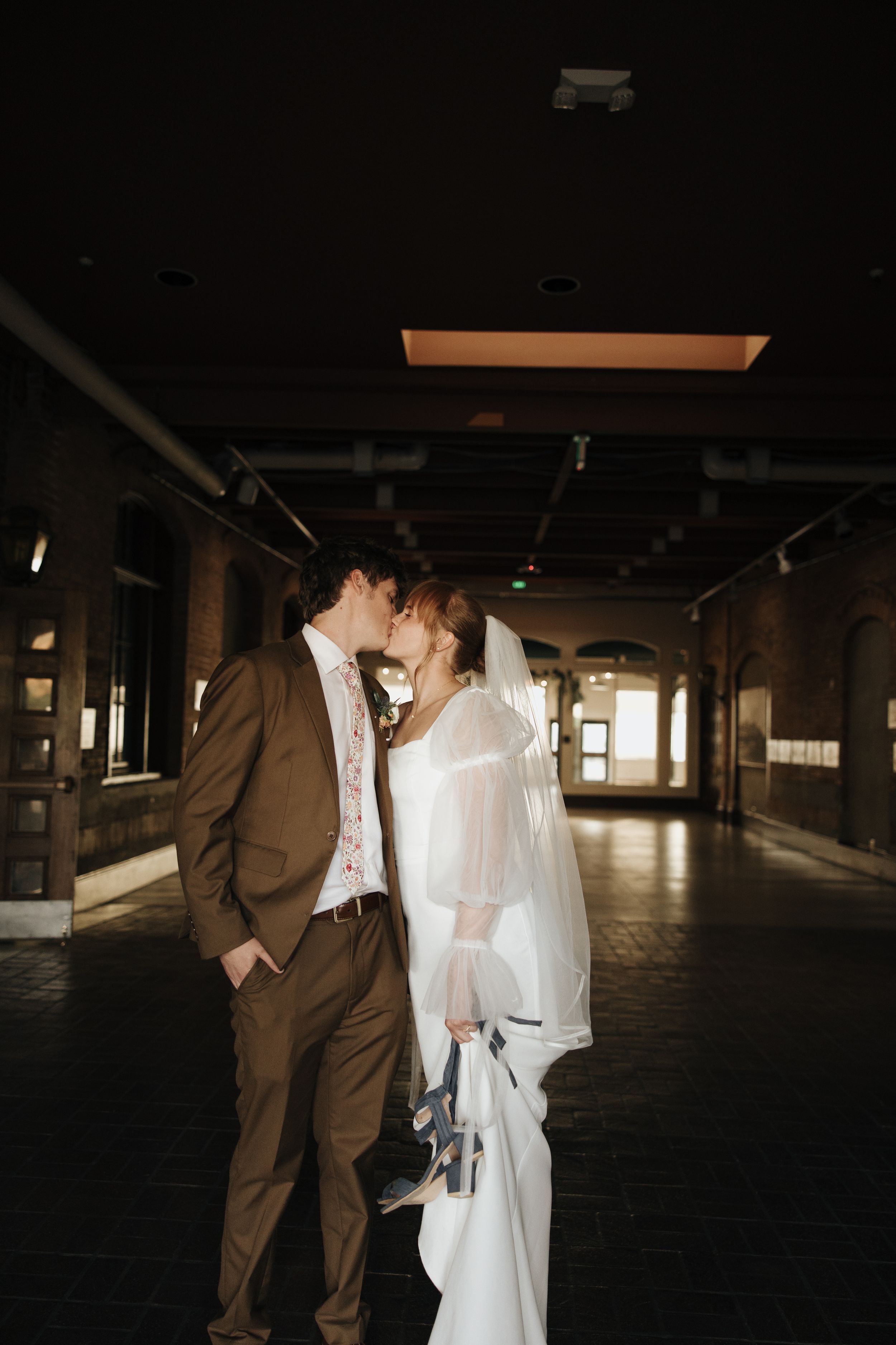 Newlyweds share intimate moment in dark industrial hallway.