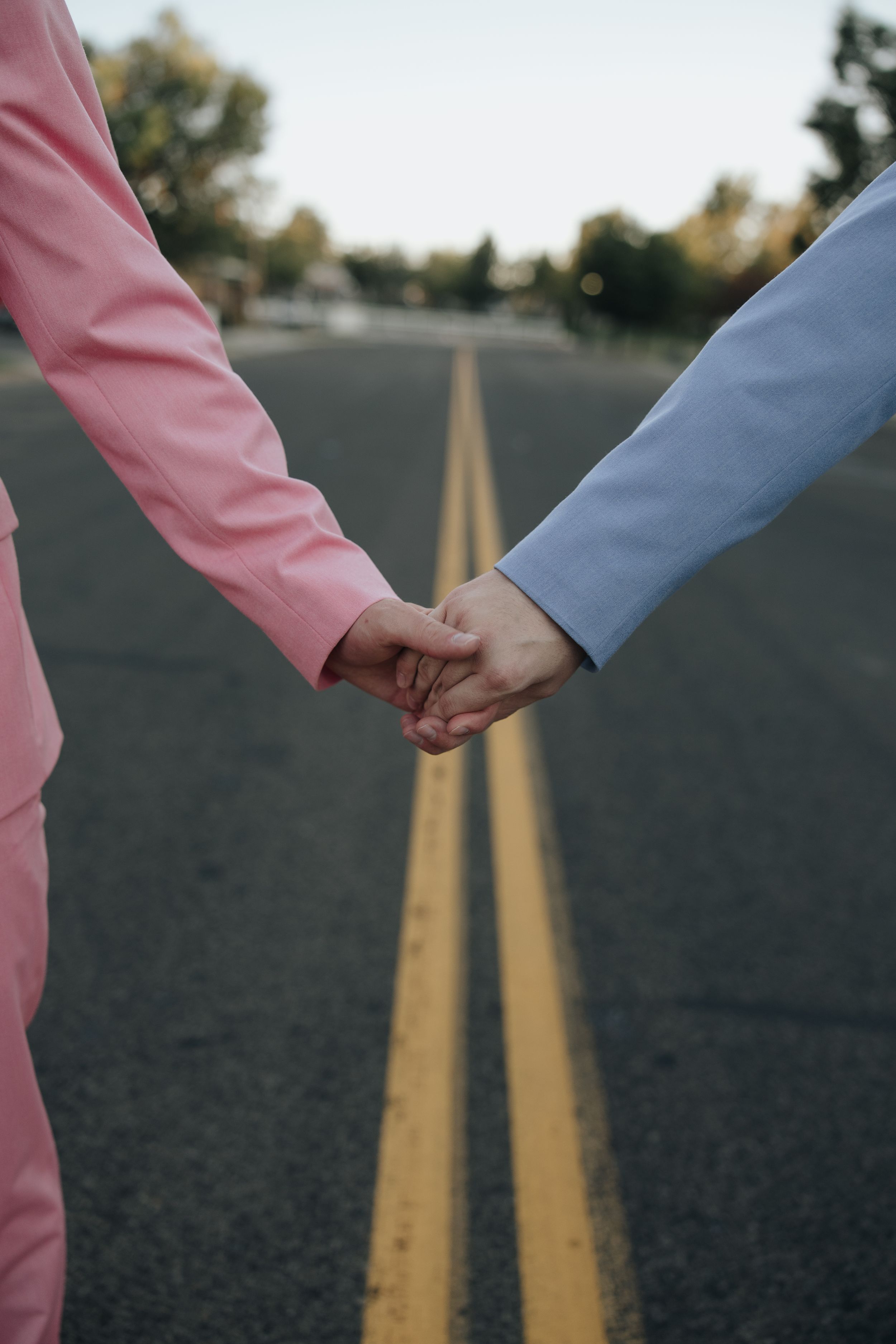 Two people holding hands stand on a road with yellow lines, one wearing pink and one wearing blue.