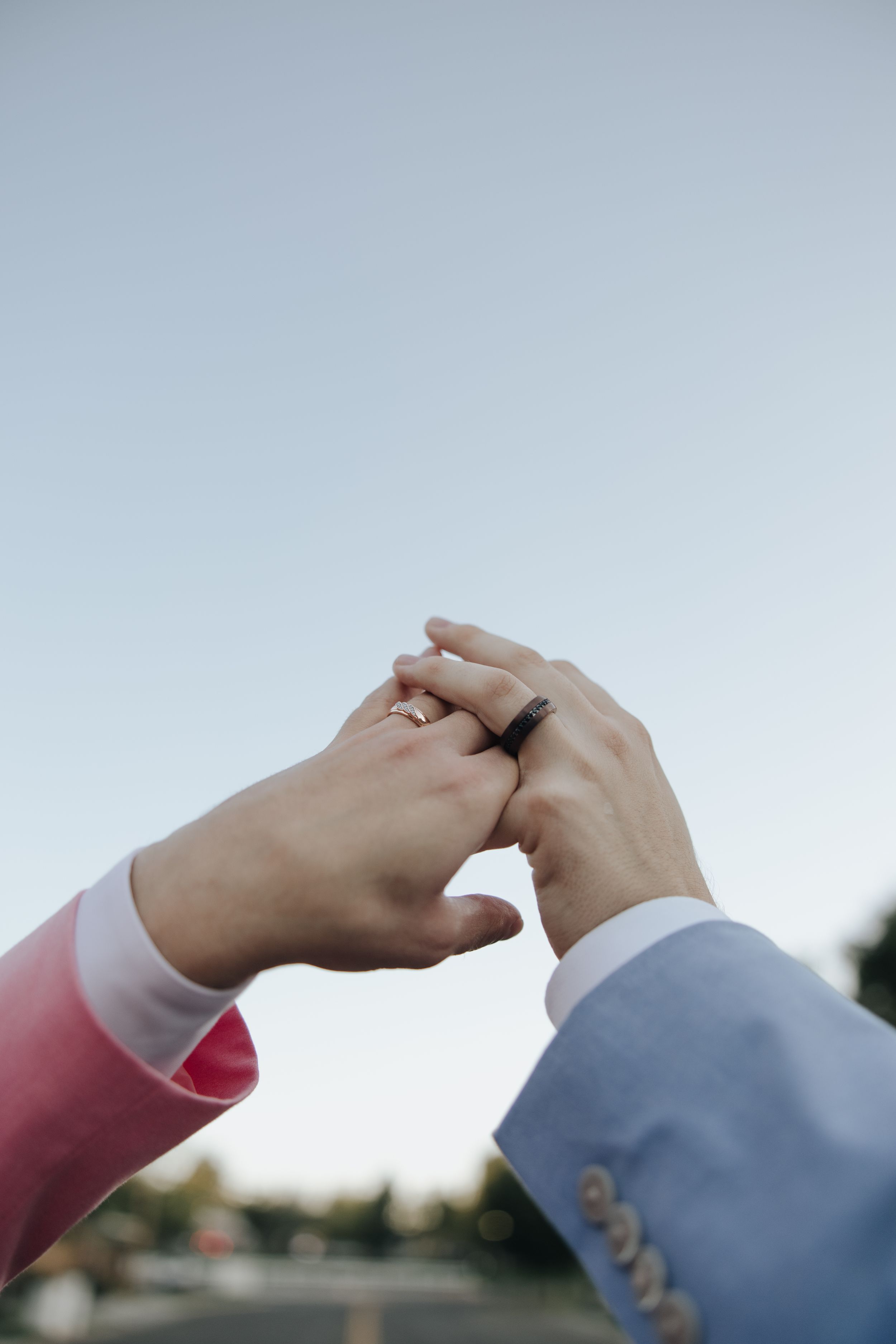 Two hands wearing formal attire join together with a ring in an intimate moment against a light sky background.