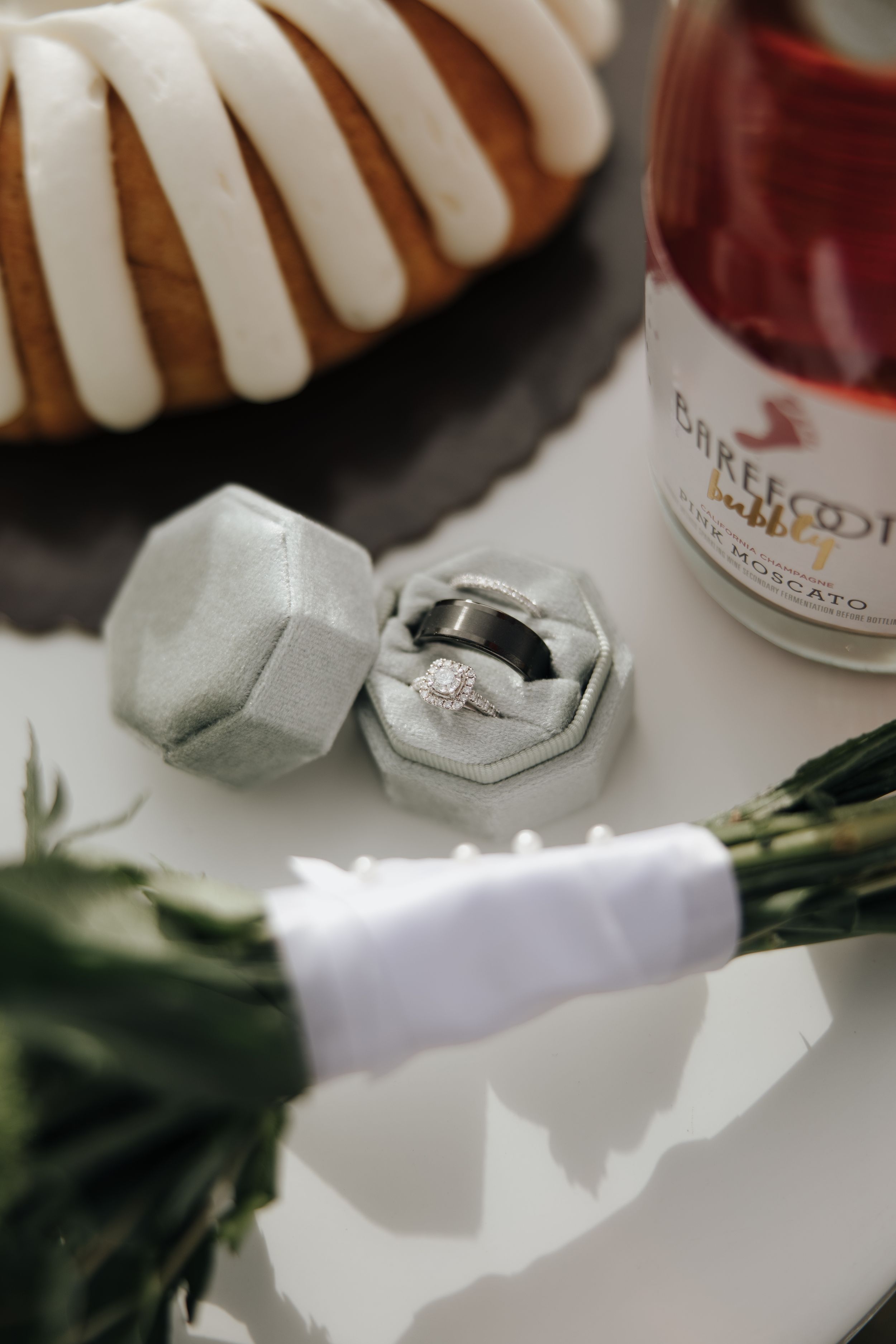 Close-up photo of engagement rings in hexagonal boxes with champagne and partial view of white floral arrangement.