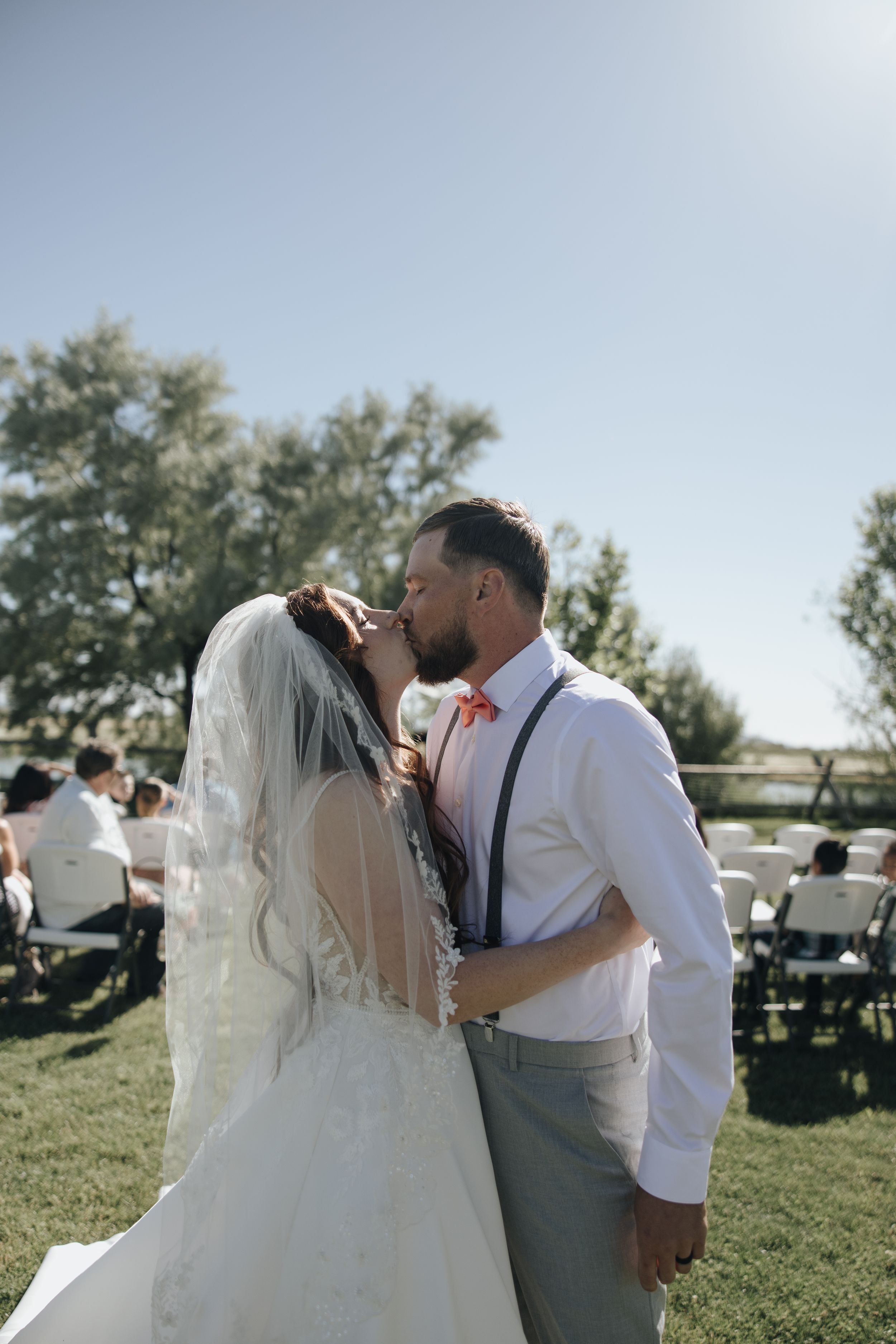 Couple shares kiss during outdoor wedding ceremony with guests seated on lawn.