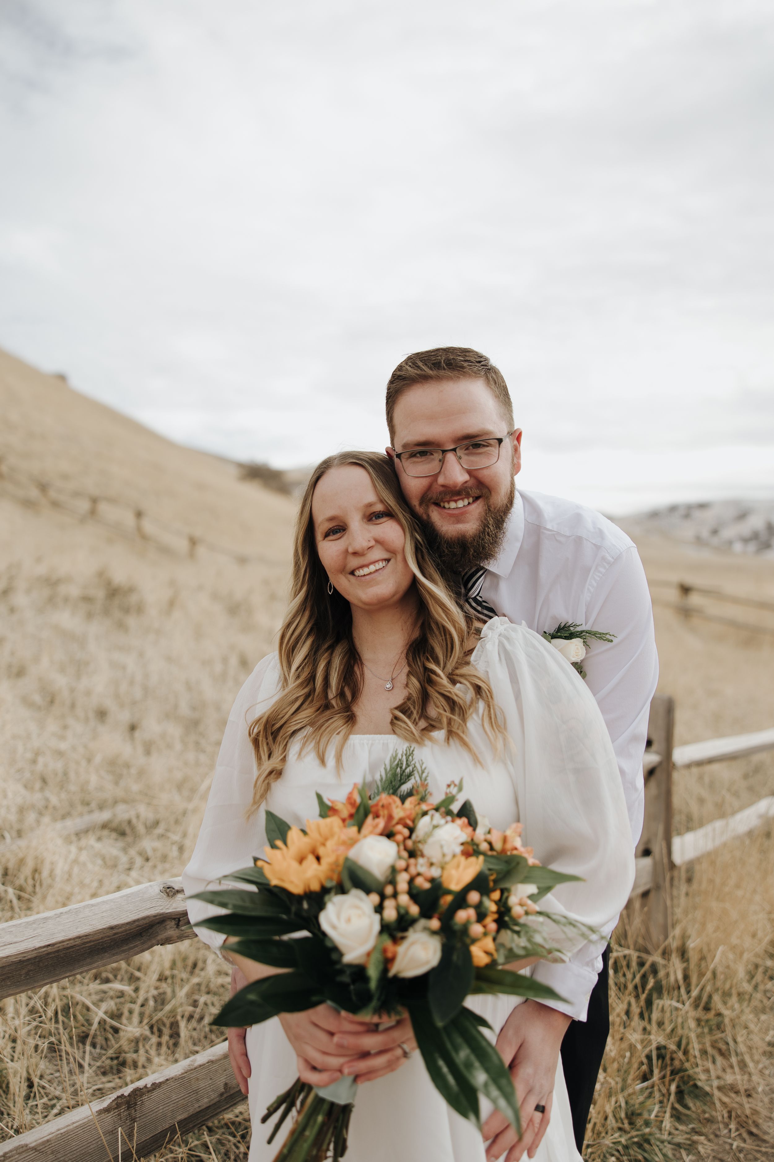 A joyful couple poses with a beautiful peach and white floral bouquet against a scenic hillside backdrop.
