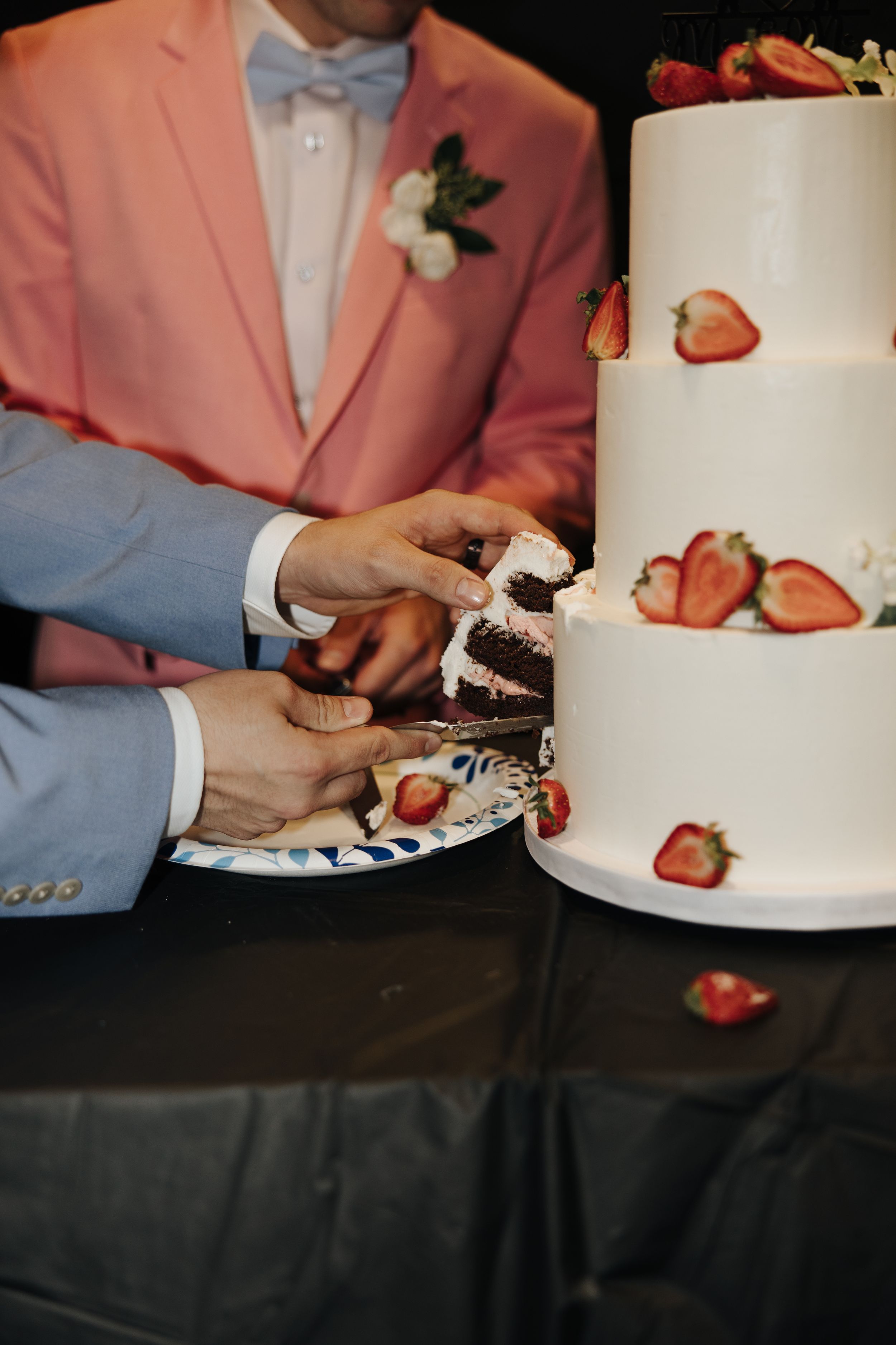 Hands cut into an elegant white wedding cake decorated with fresh strawberries as a pink suit-clad figure stands nearby.