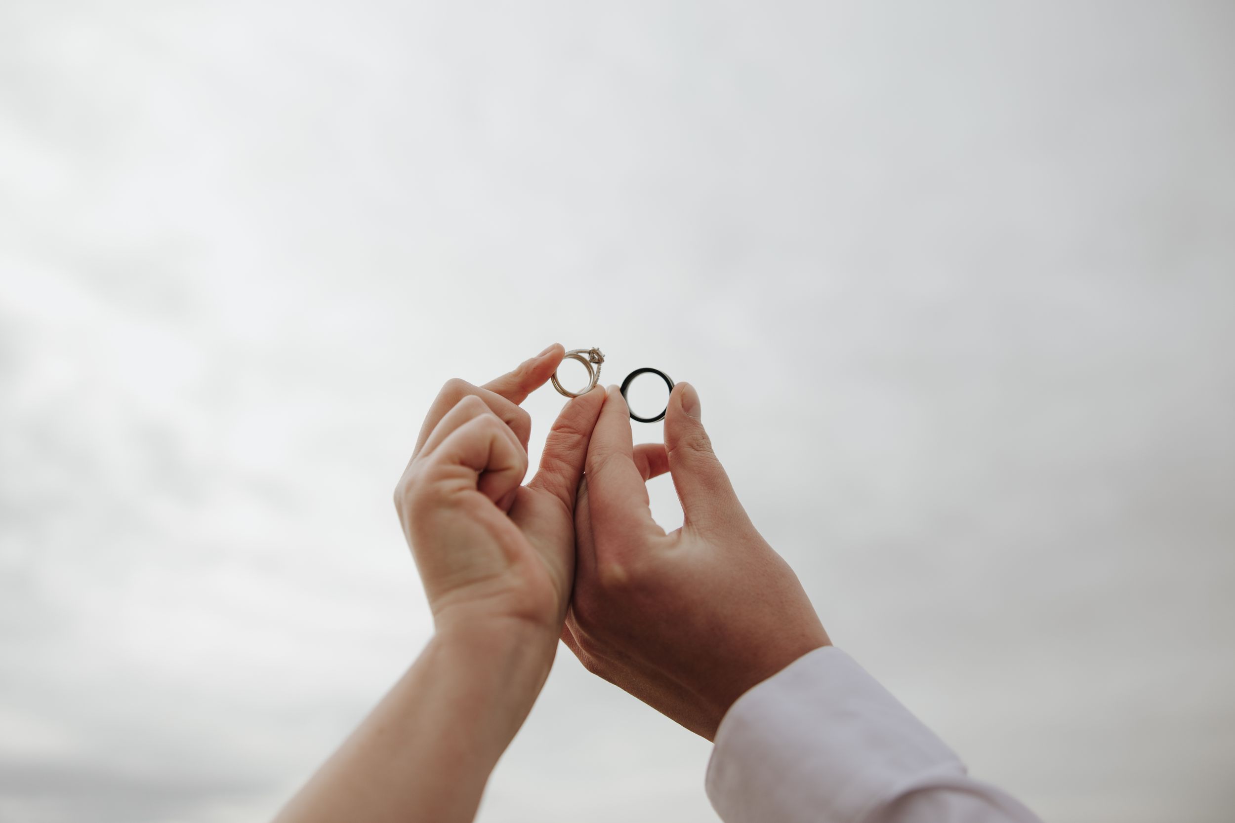 Hands holding silver ring against cloudy sky background during proposal moment at sunset.