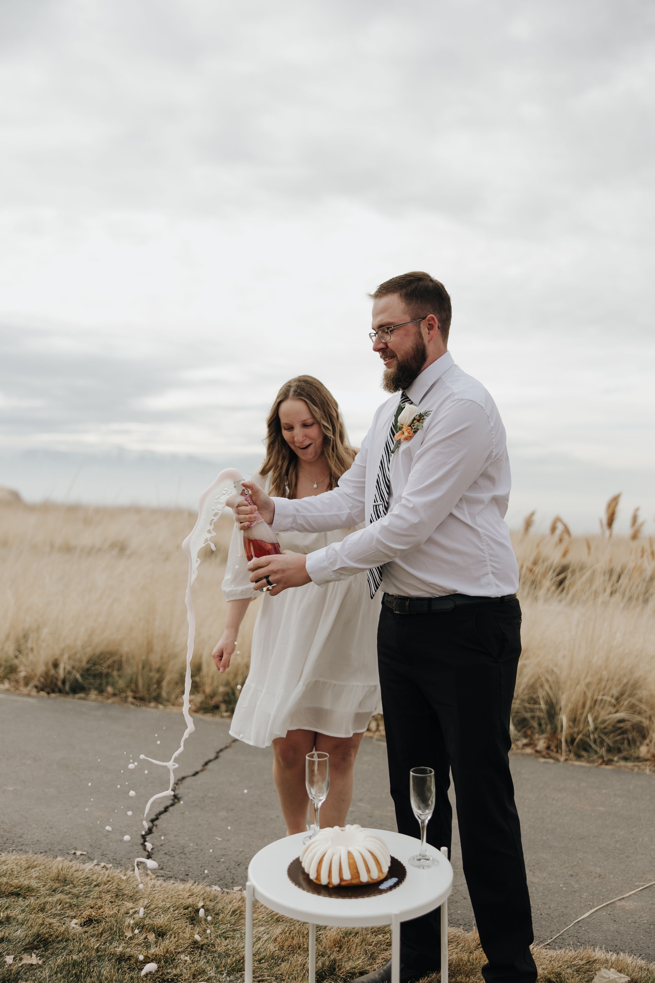 A couple celebrates by popping champagne over a bundt cake at their outdoor wedding reception.