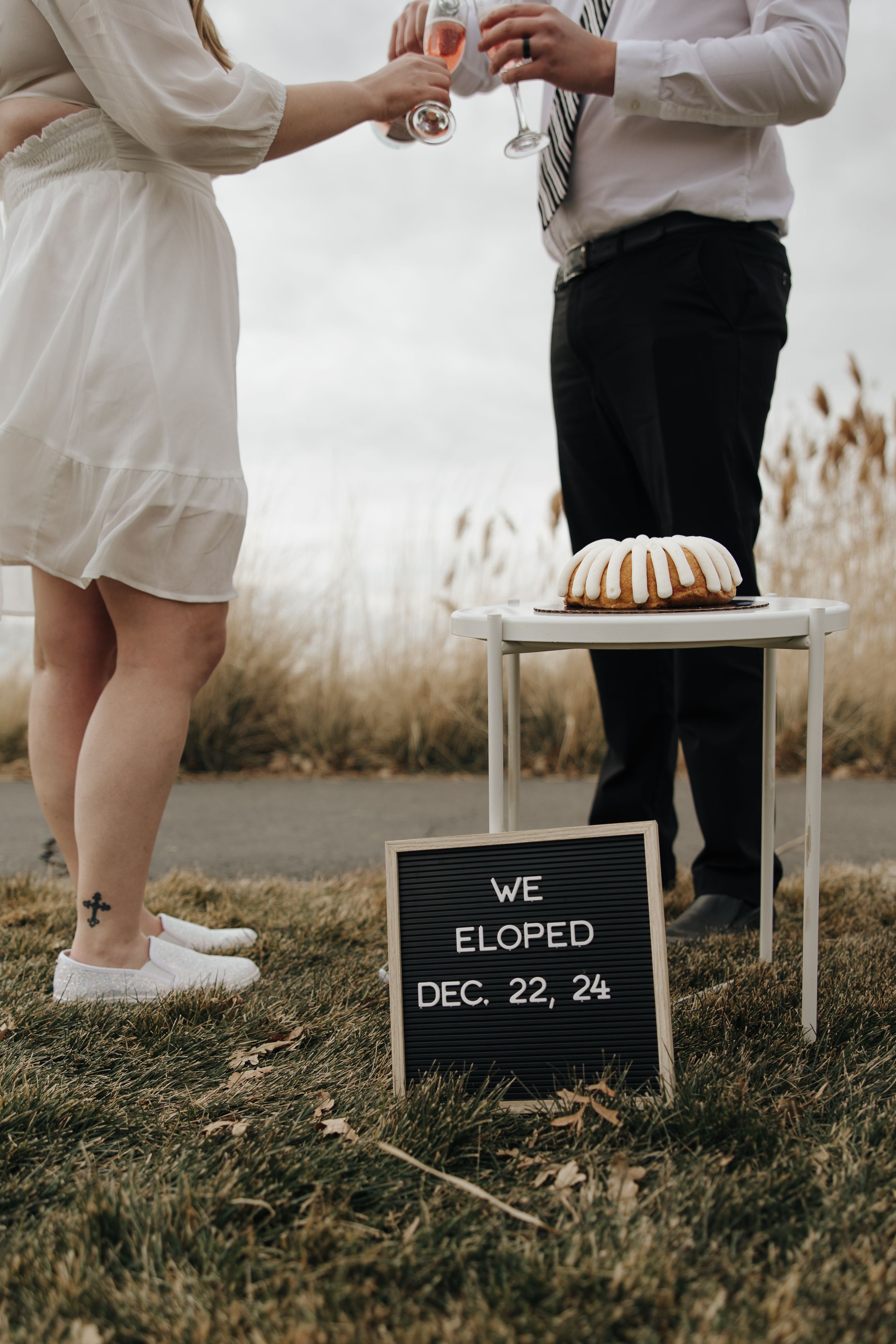 Closeup of champagne toast with couple's feet and bundt cake on white table with elopement sign.