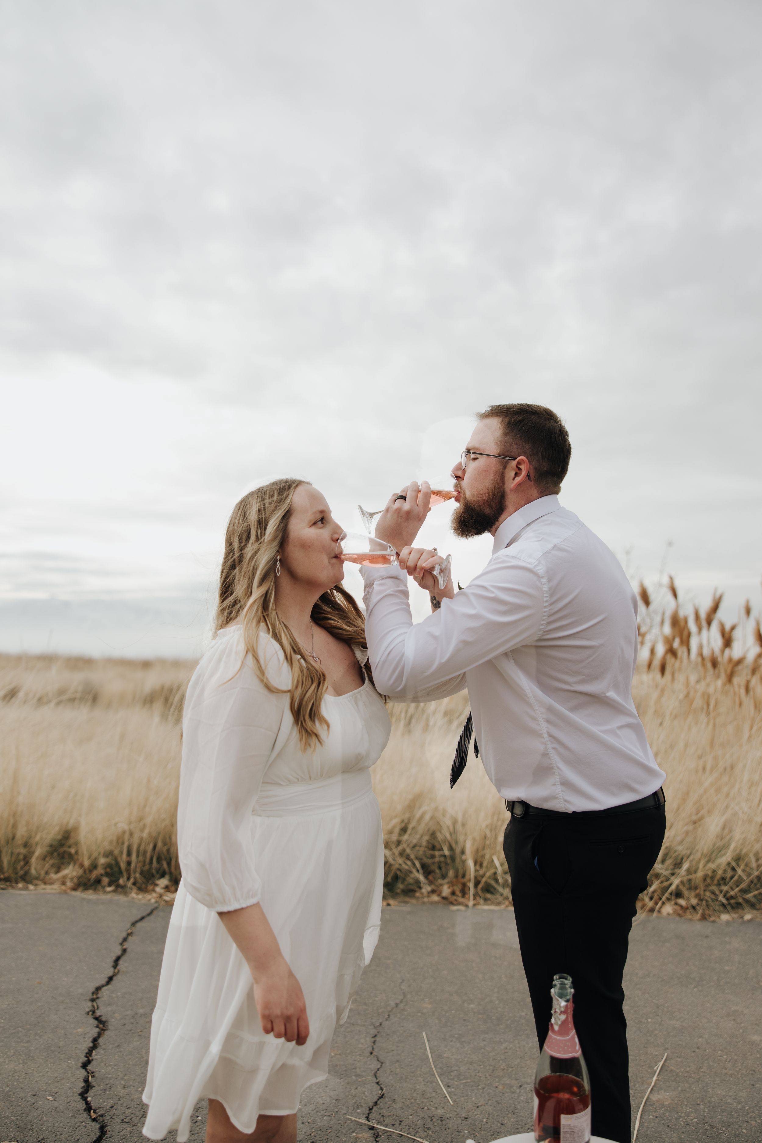 A couple in white clothing shares a playful moment drinking wine together in a field with tall grass on an overcast day.