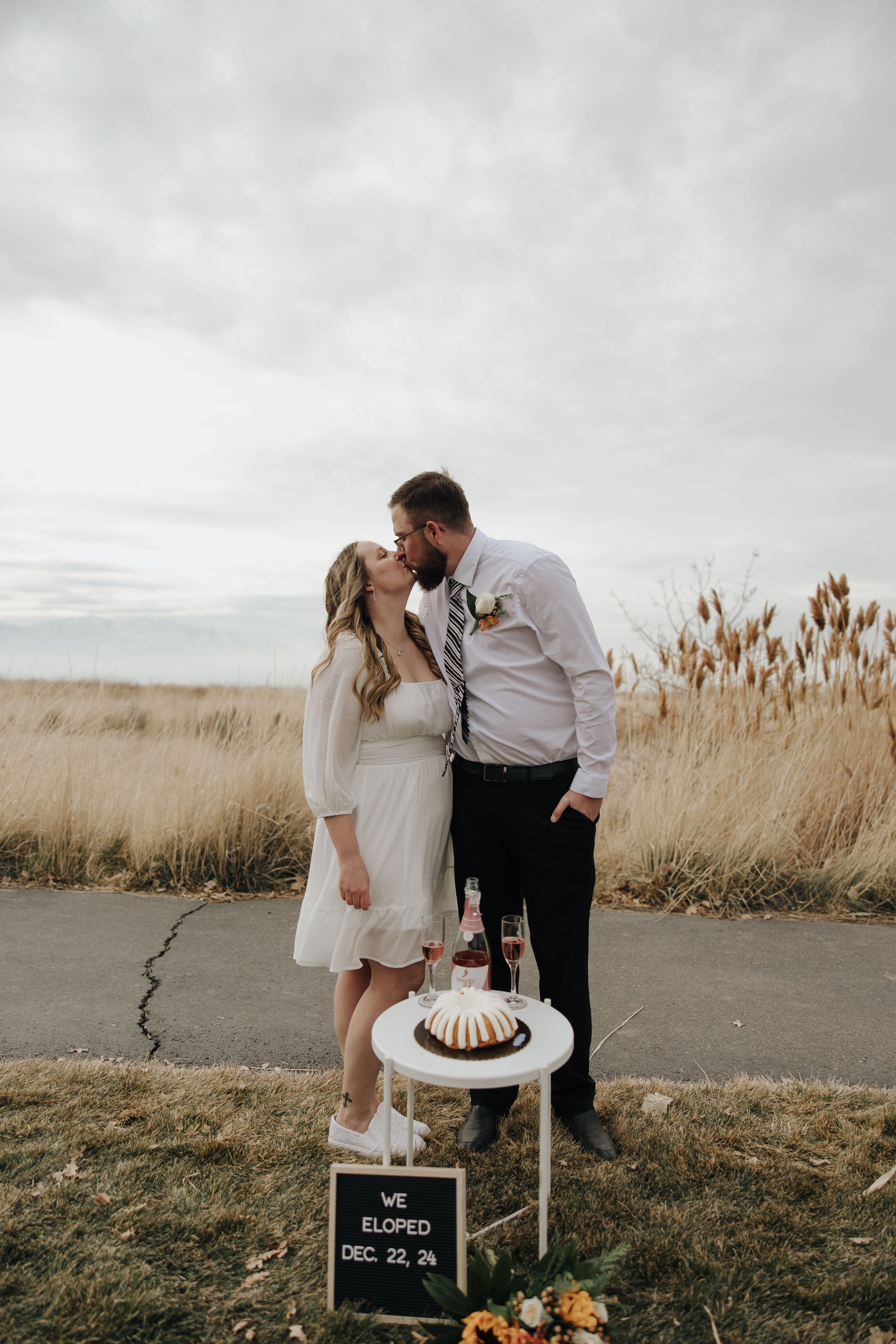 Romantic elopement scene with couple sharing intimate moment over cake and champagne at sunset.