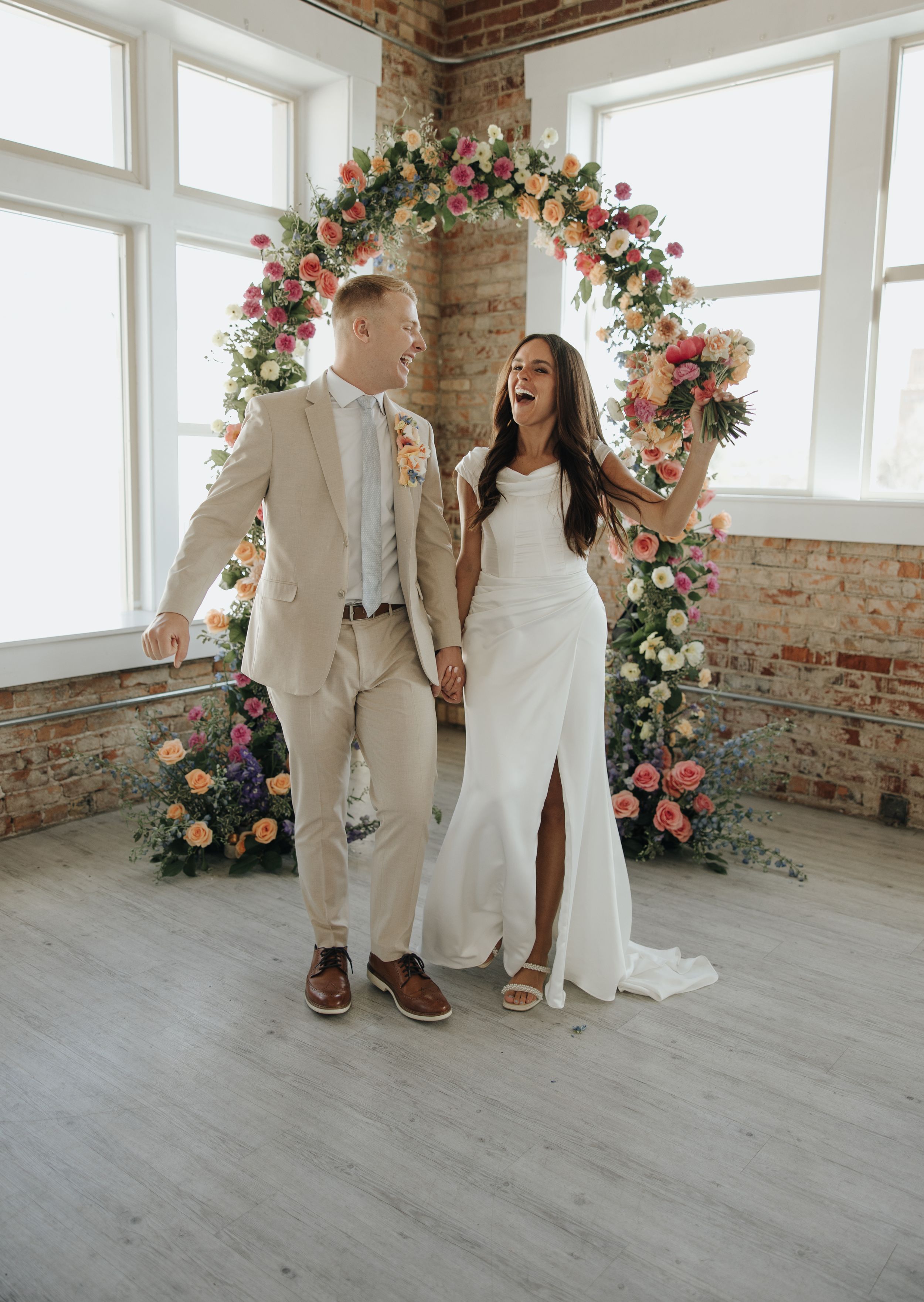Joyful wedding couple laughs under floral arch with pink and peach blooms in sunlit room.