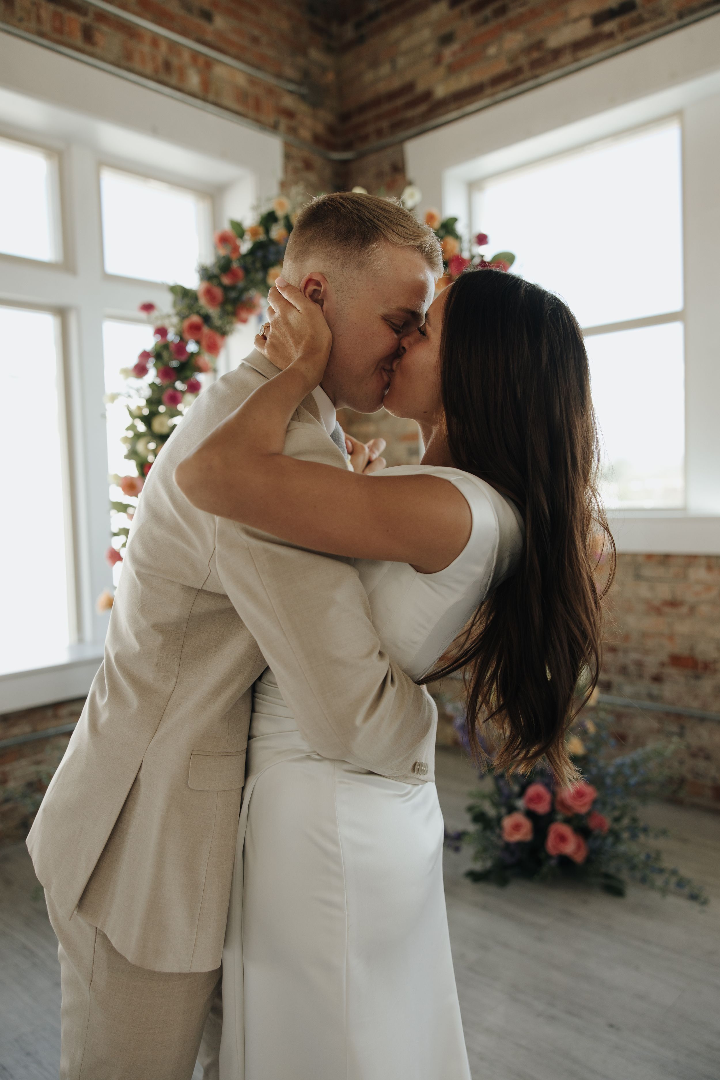 Intimate wedding moment captured against windows with floral arch decor in rustic brick room.