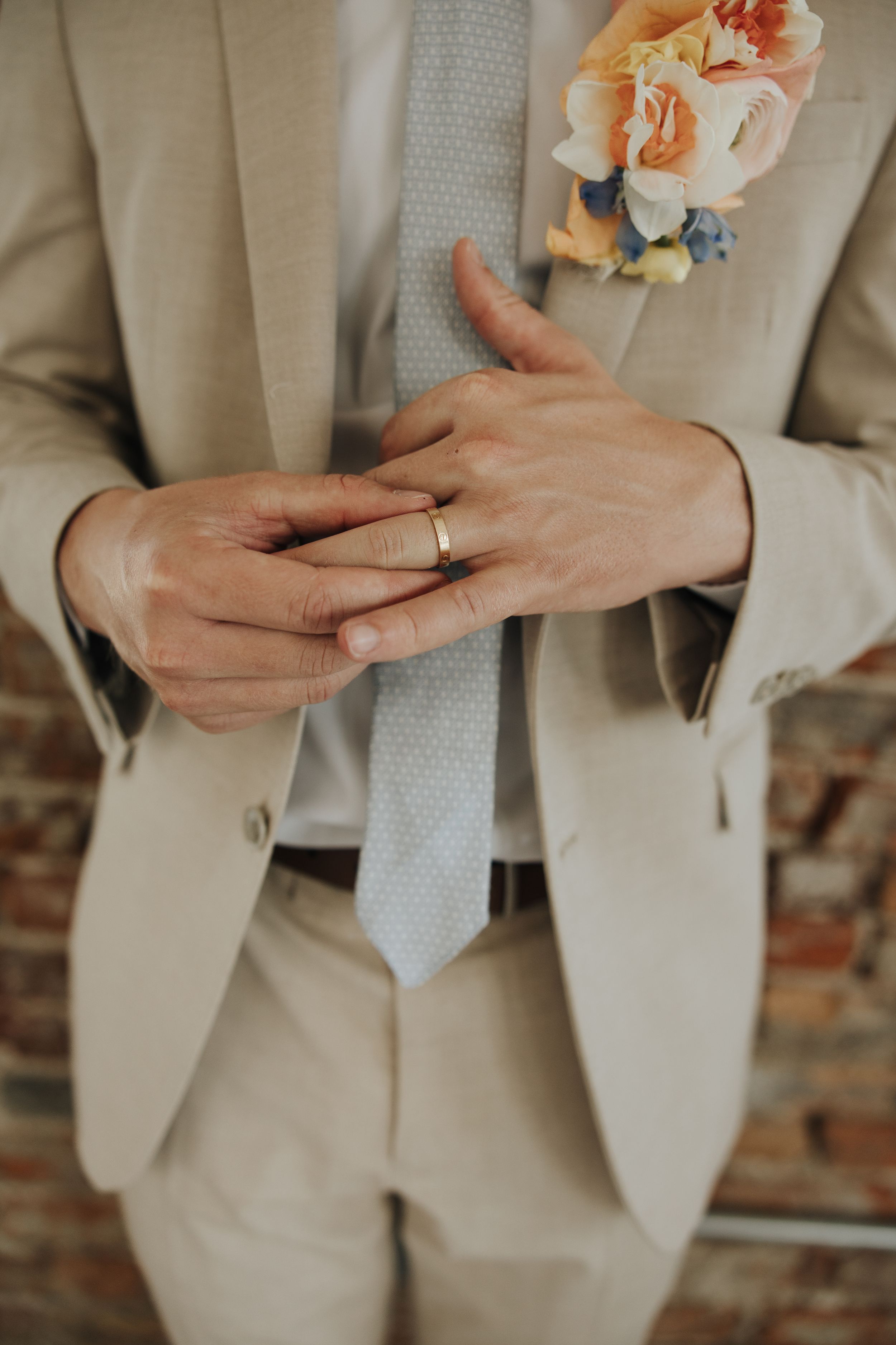 Detail shot of hands adjusting a wedding ring with a boutonniere on a beige suit.