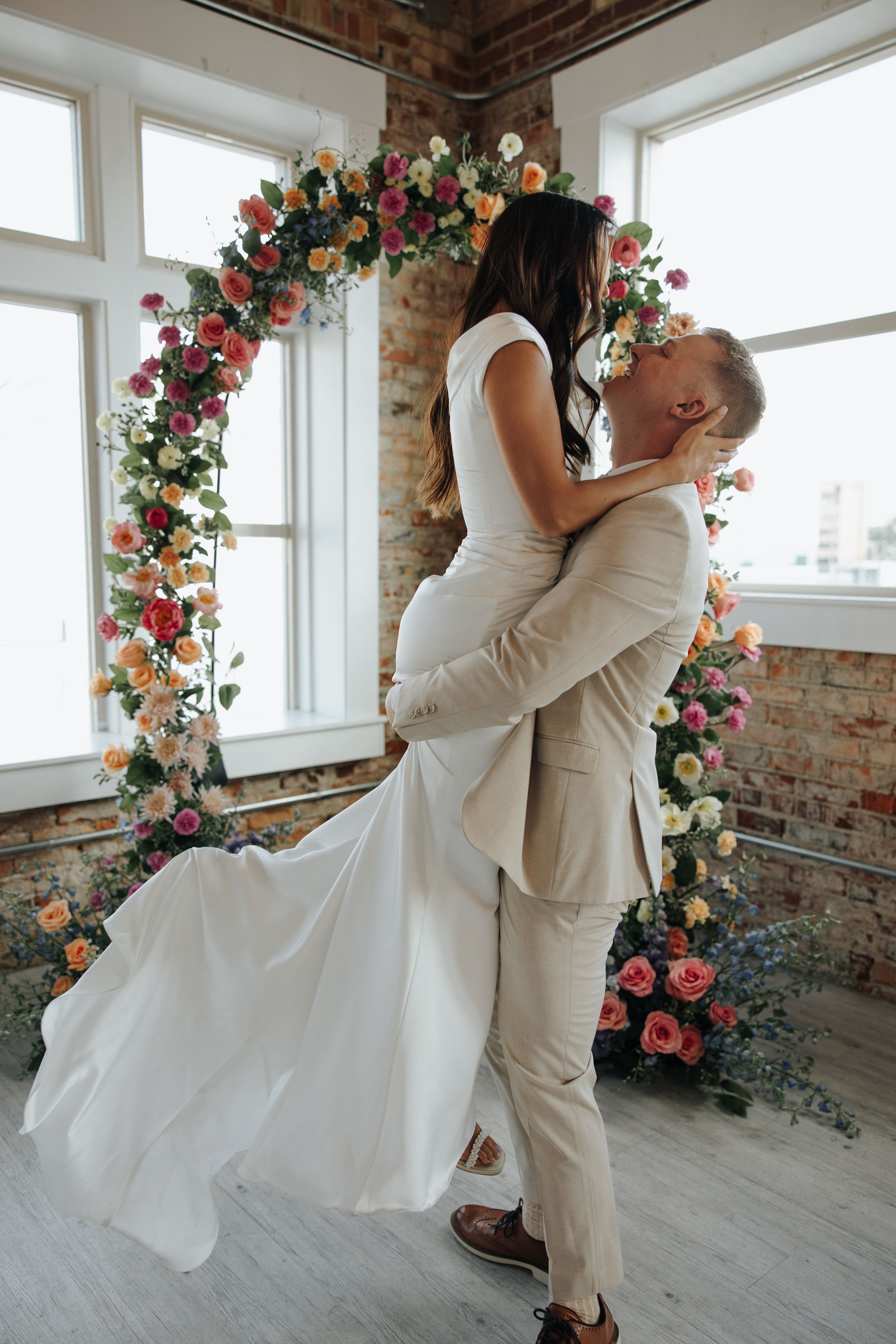 Romantic lift pose under a floral arch with pink and peach blooms against brick walls.