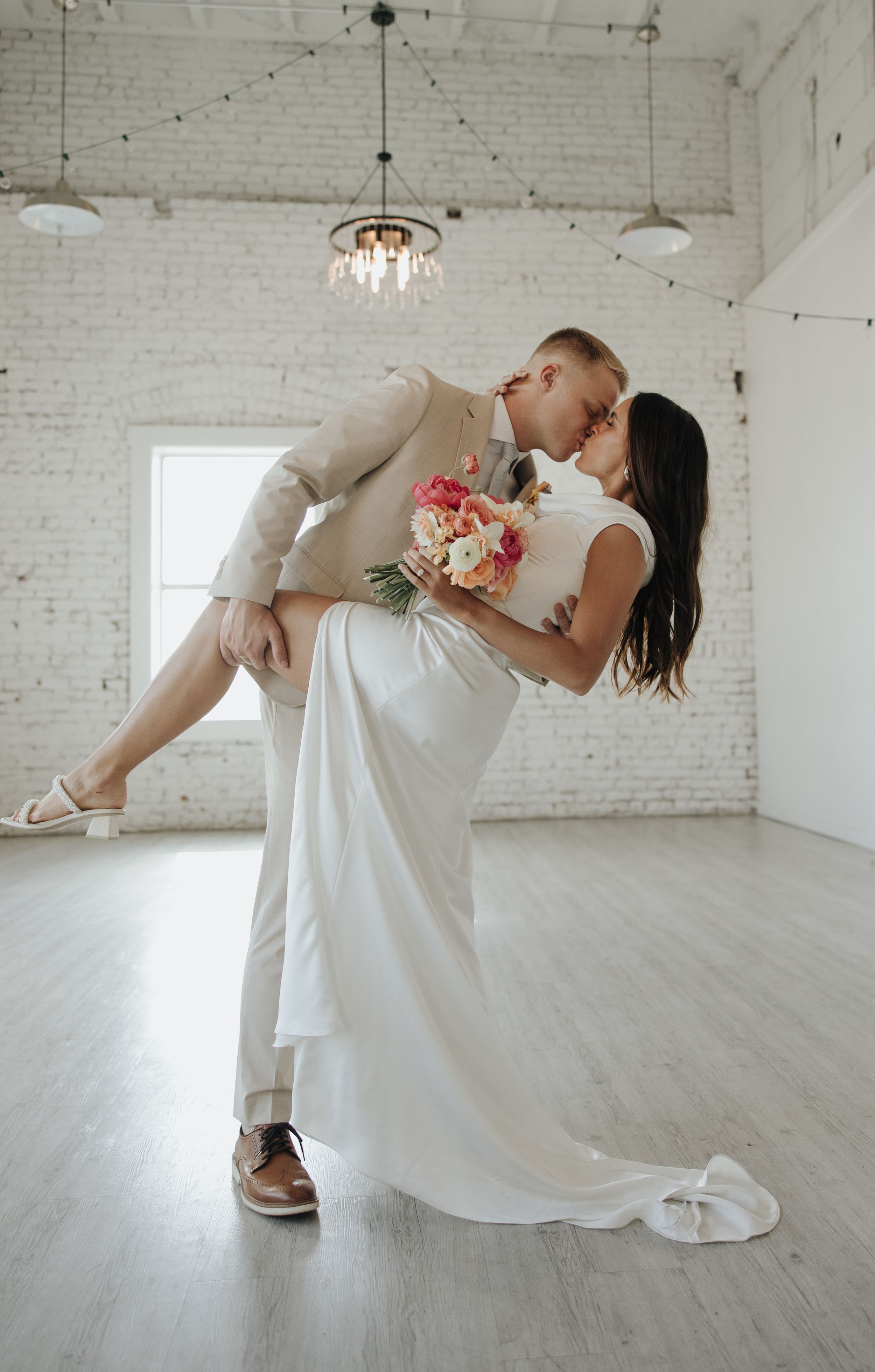 Romantic dip pose in a white industrial venue with string lights and brick walls.