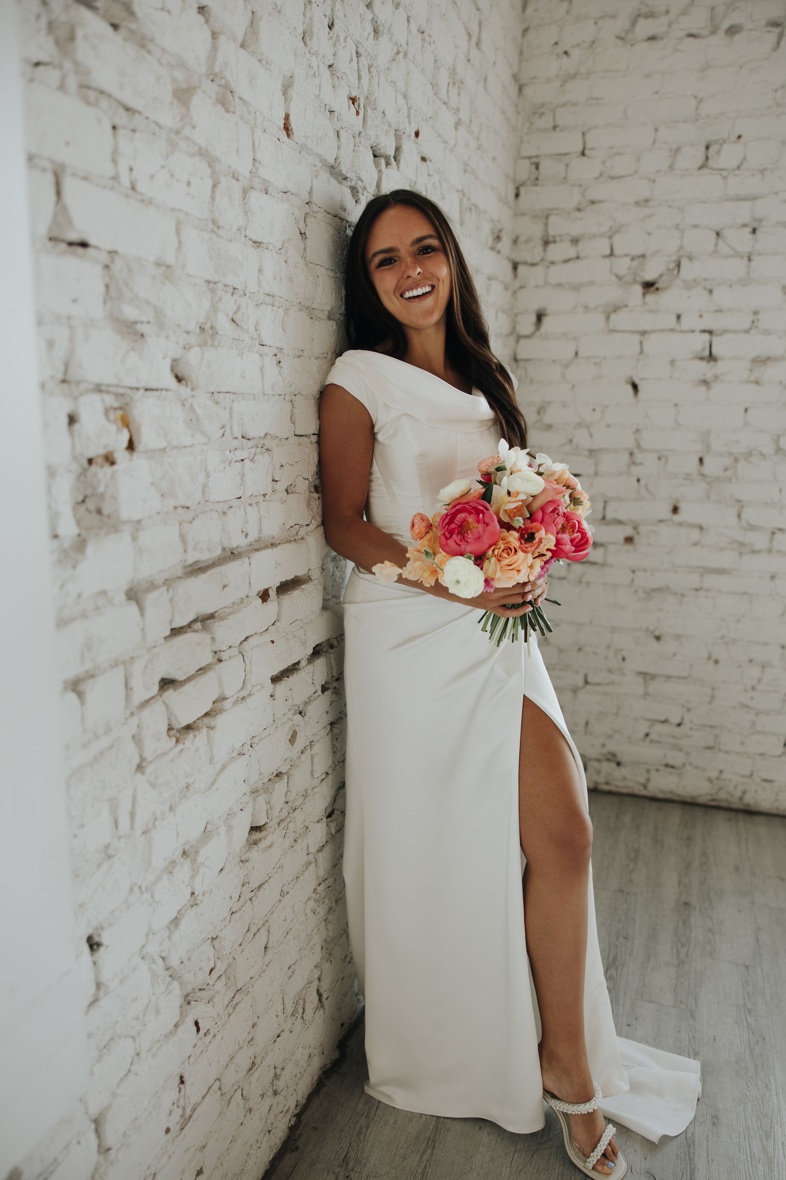Bride in elegant white dress with thigh slit poses with pink bouquet against white brick wall.