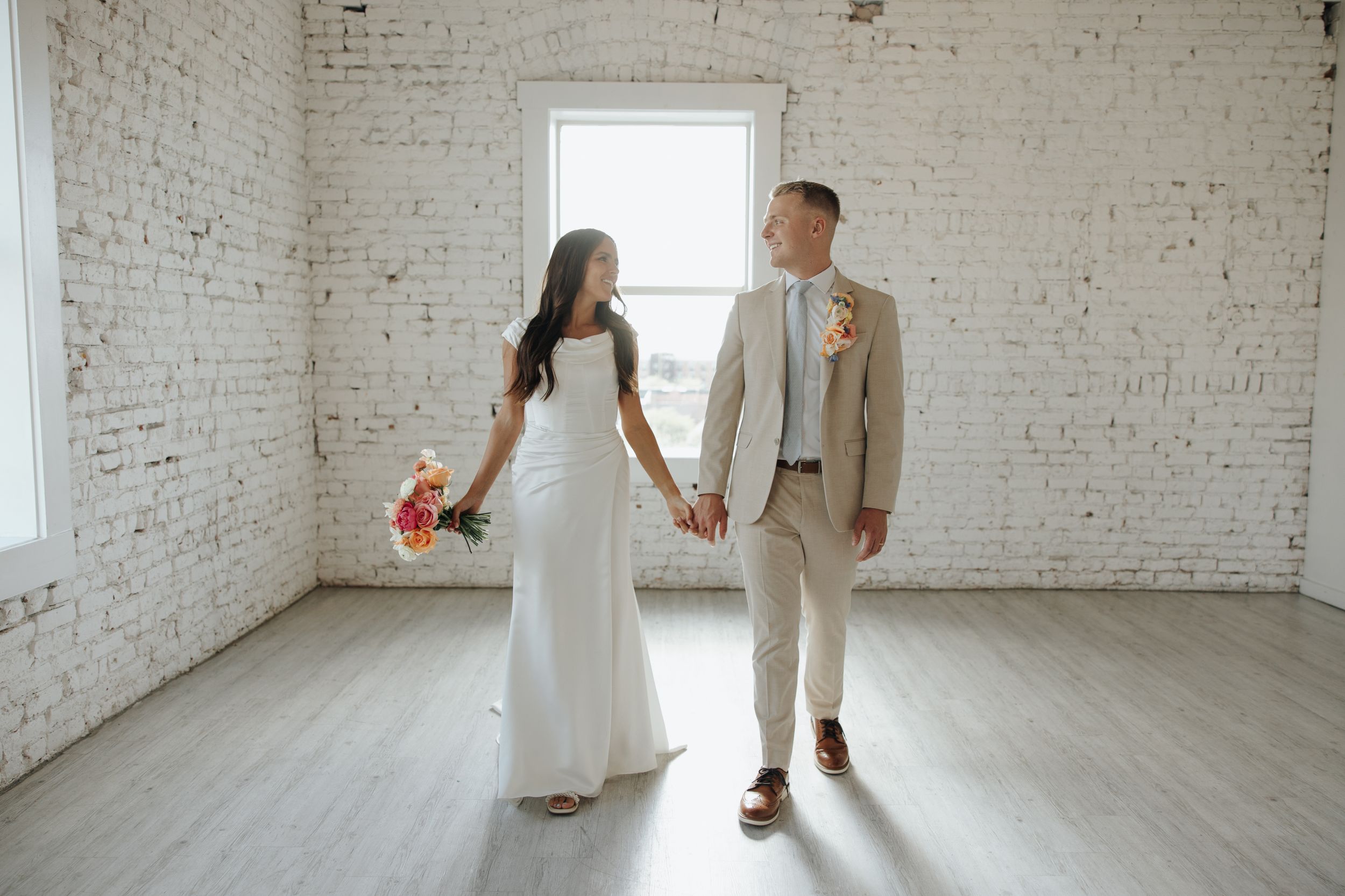 A wedding couple holds hands in an elegant white brick room with natural lighting streaming through the windows.