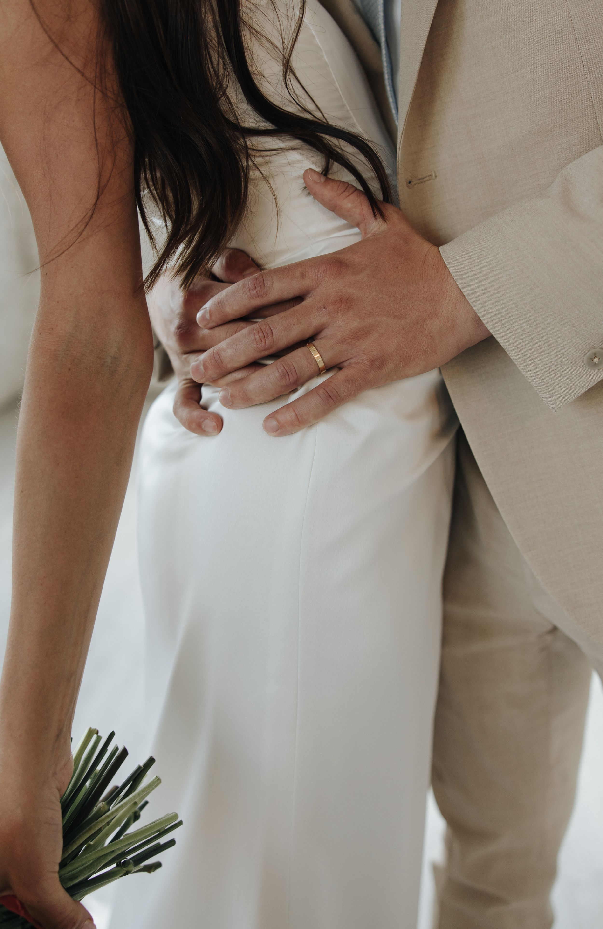Close-up of hands embracing around a bride's waist in a white dress against beige suit.