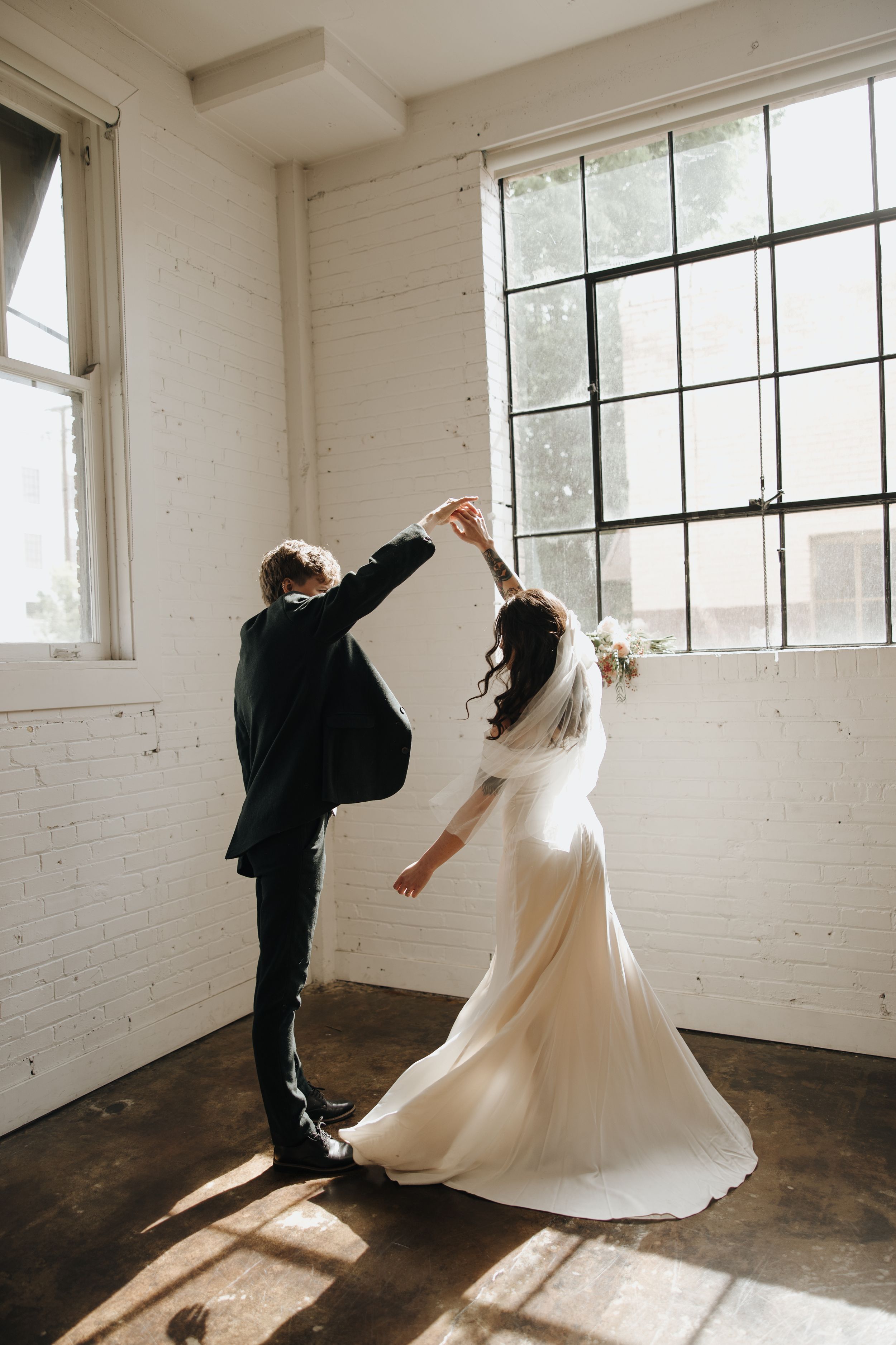Dancing couple in wedding attire twirling in sunlit room with large windows.