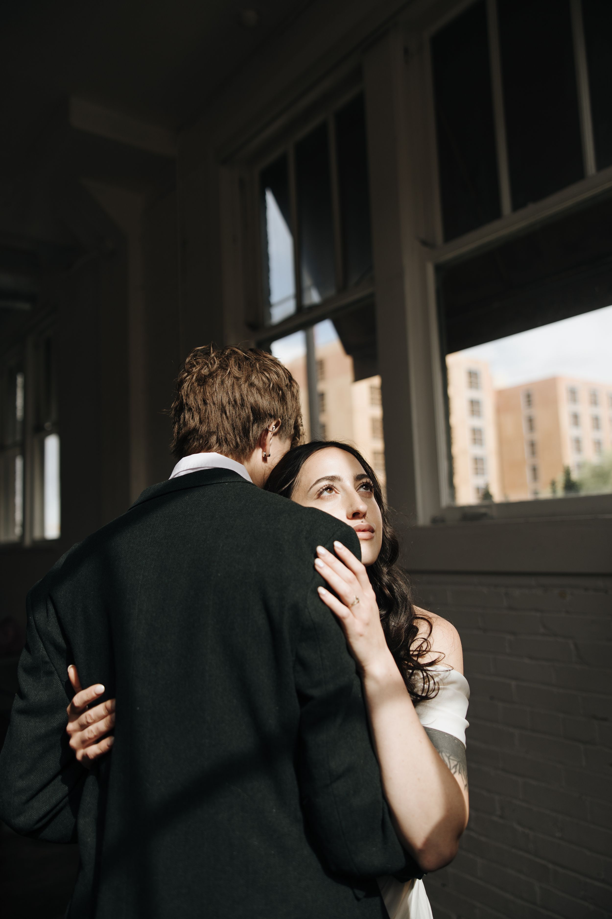 Intimate moment between couple sharing an embrace near windows in a dark indoor setting.
