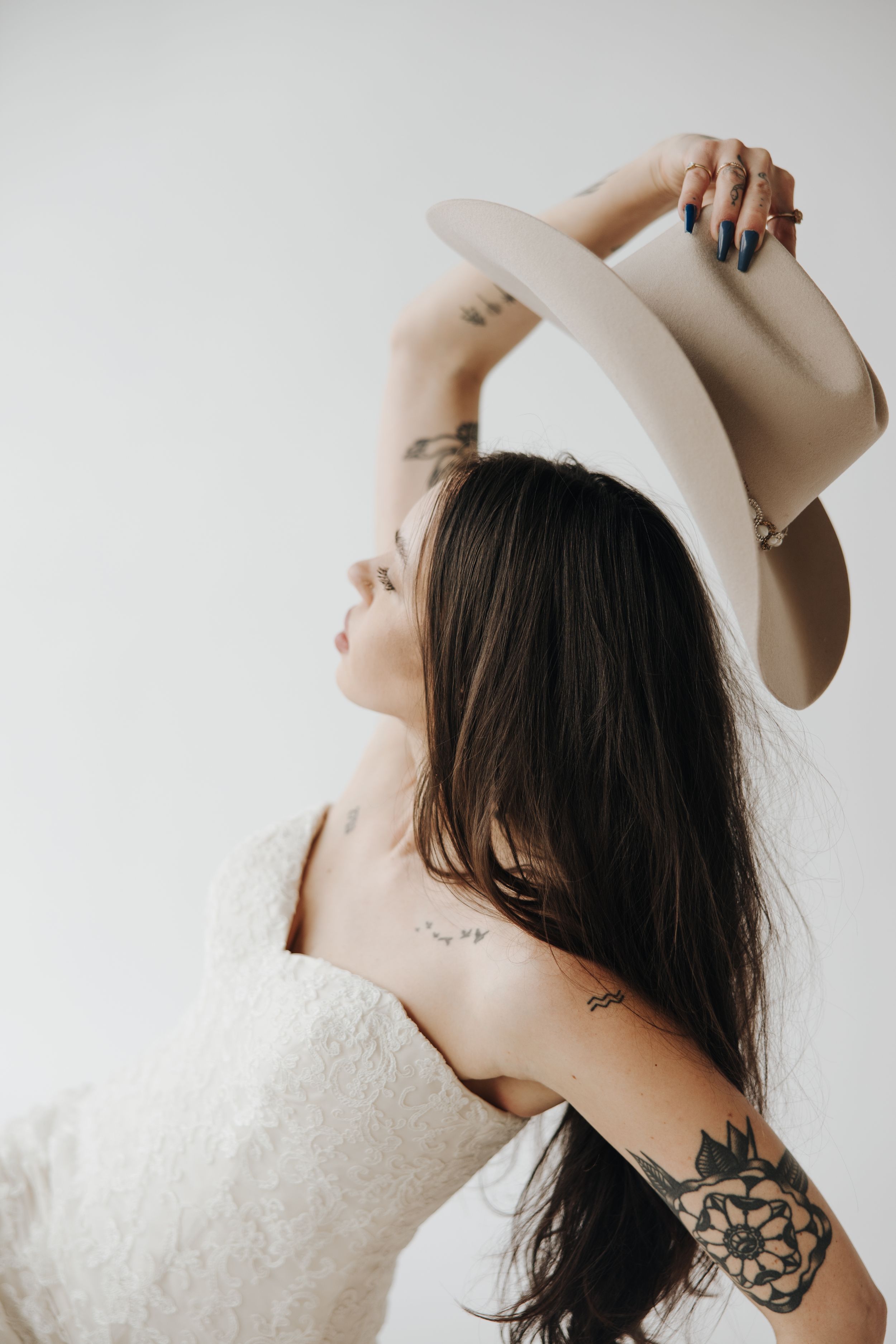 Woman in white dress poses with cream cowboy hat showing tattoos against plain backdrop.