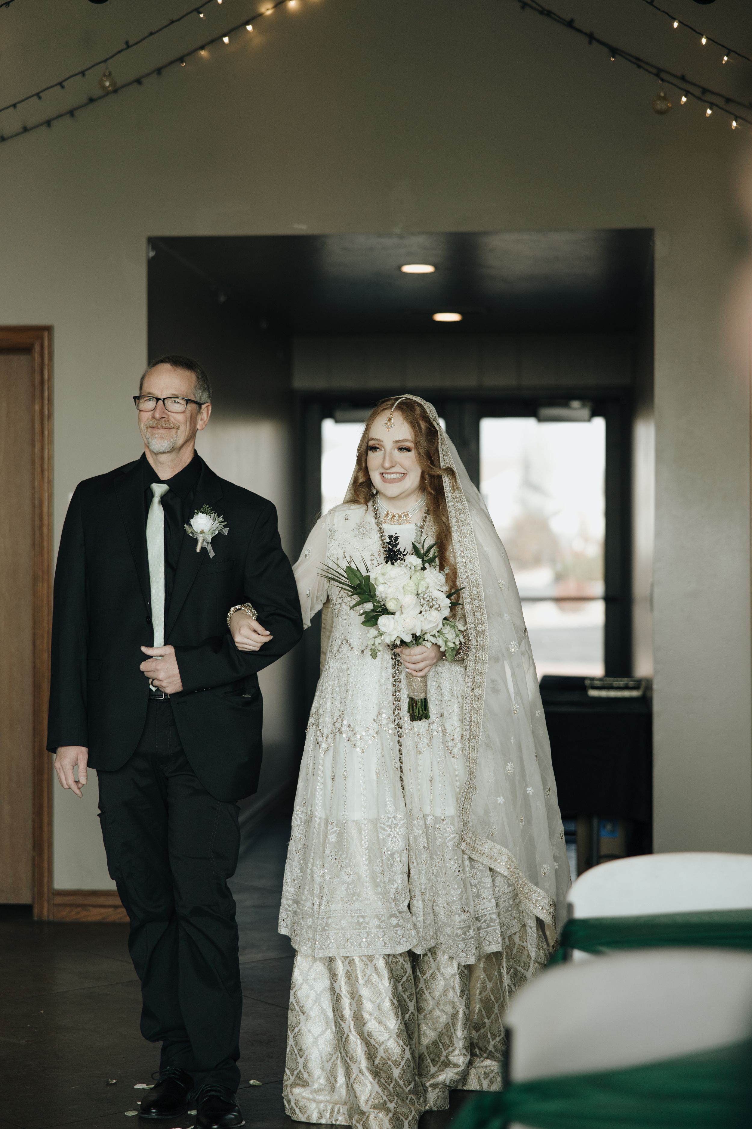 An elegant winter wedding processional with a couple walking down the aisle in formal attire and delicate string lights overhead.