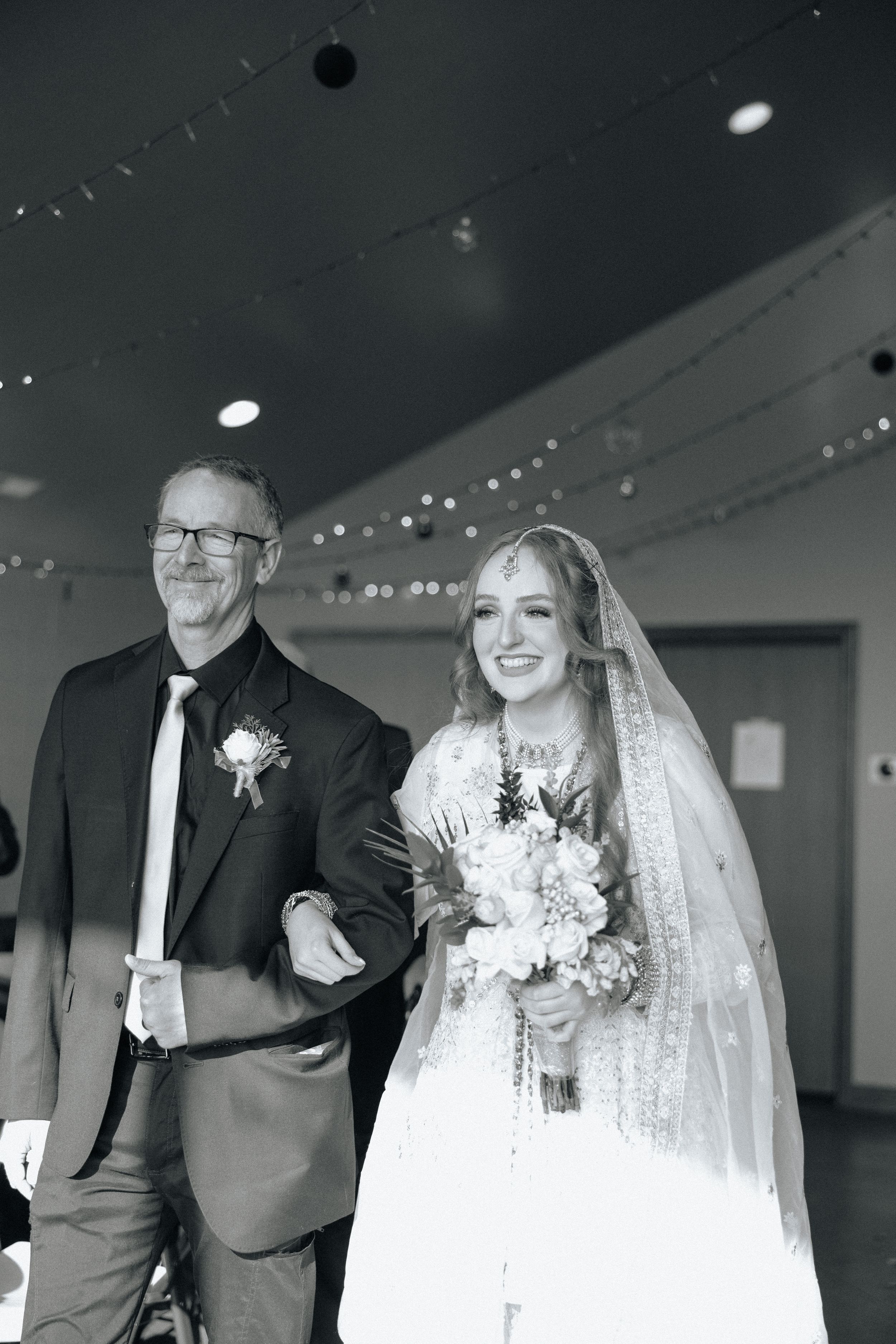 A black and white wedding photo showing two people walking arm in arm down an aisle under string lights indoors.