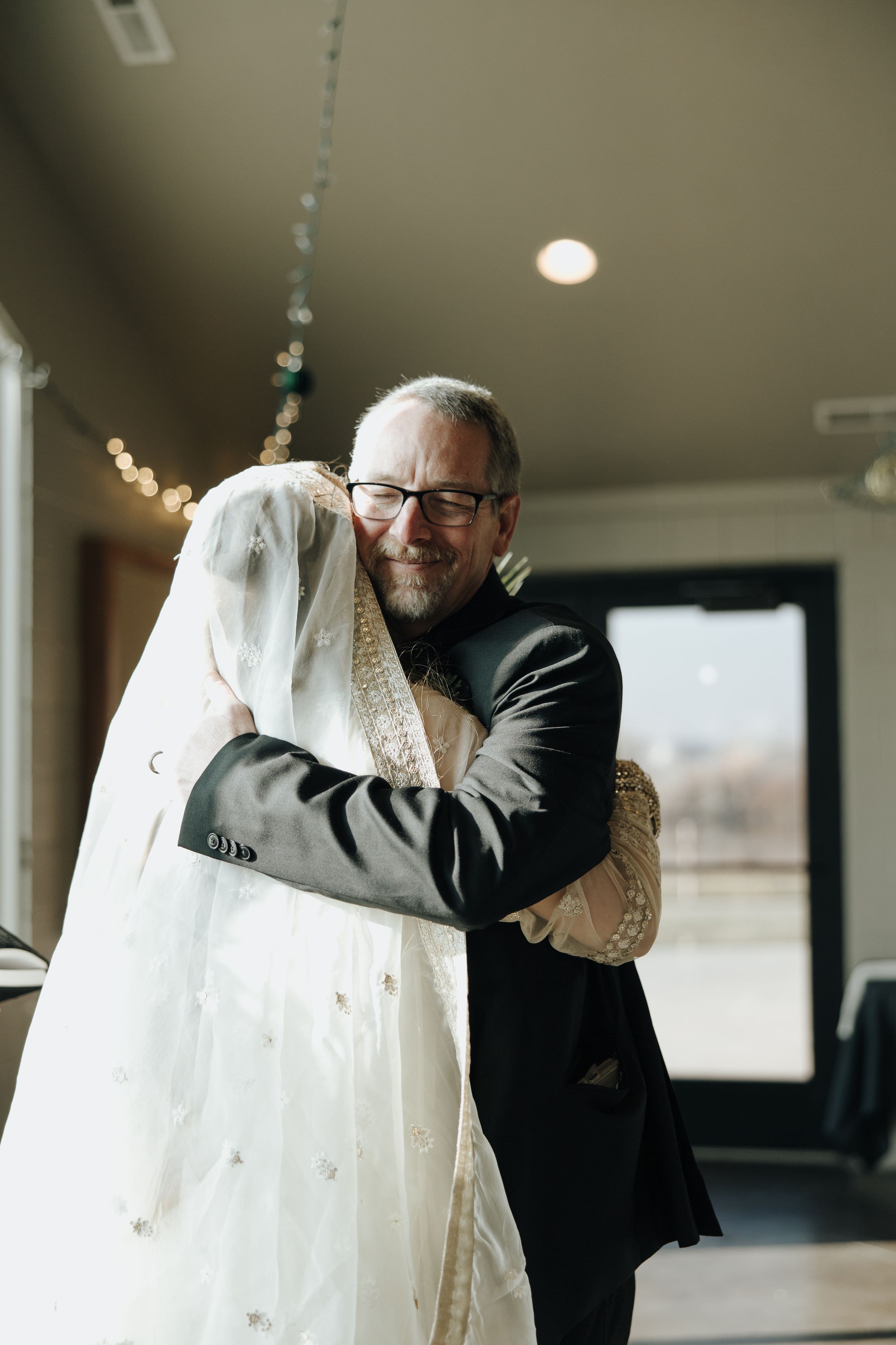 A person in a dark suit hugs a white wedding dress on a hanger while standing in a room with string lights.