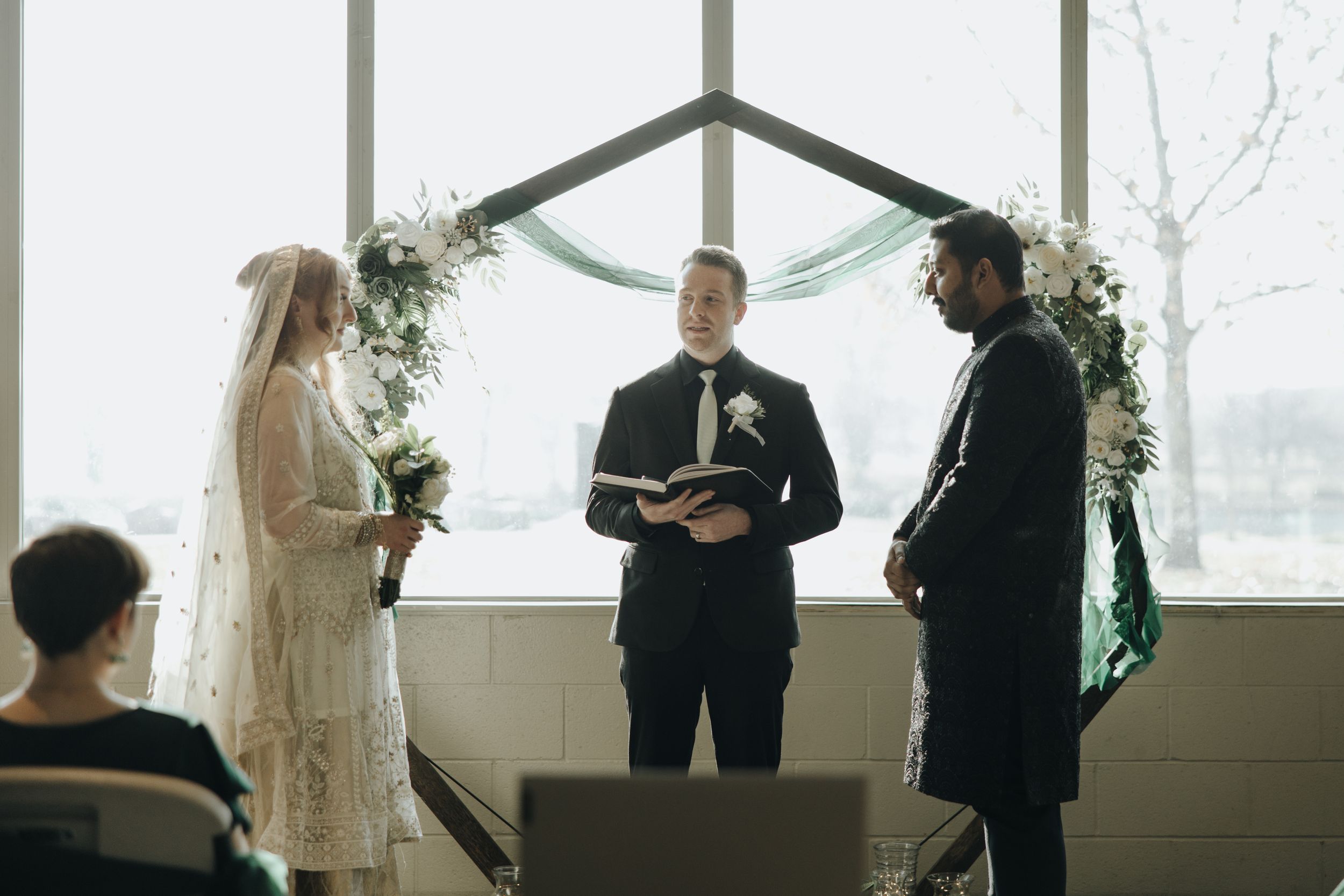 A wedding ceremony takes place under a draped arch by a window with a couple being married by an officiant.