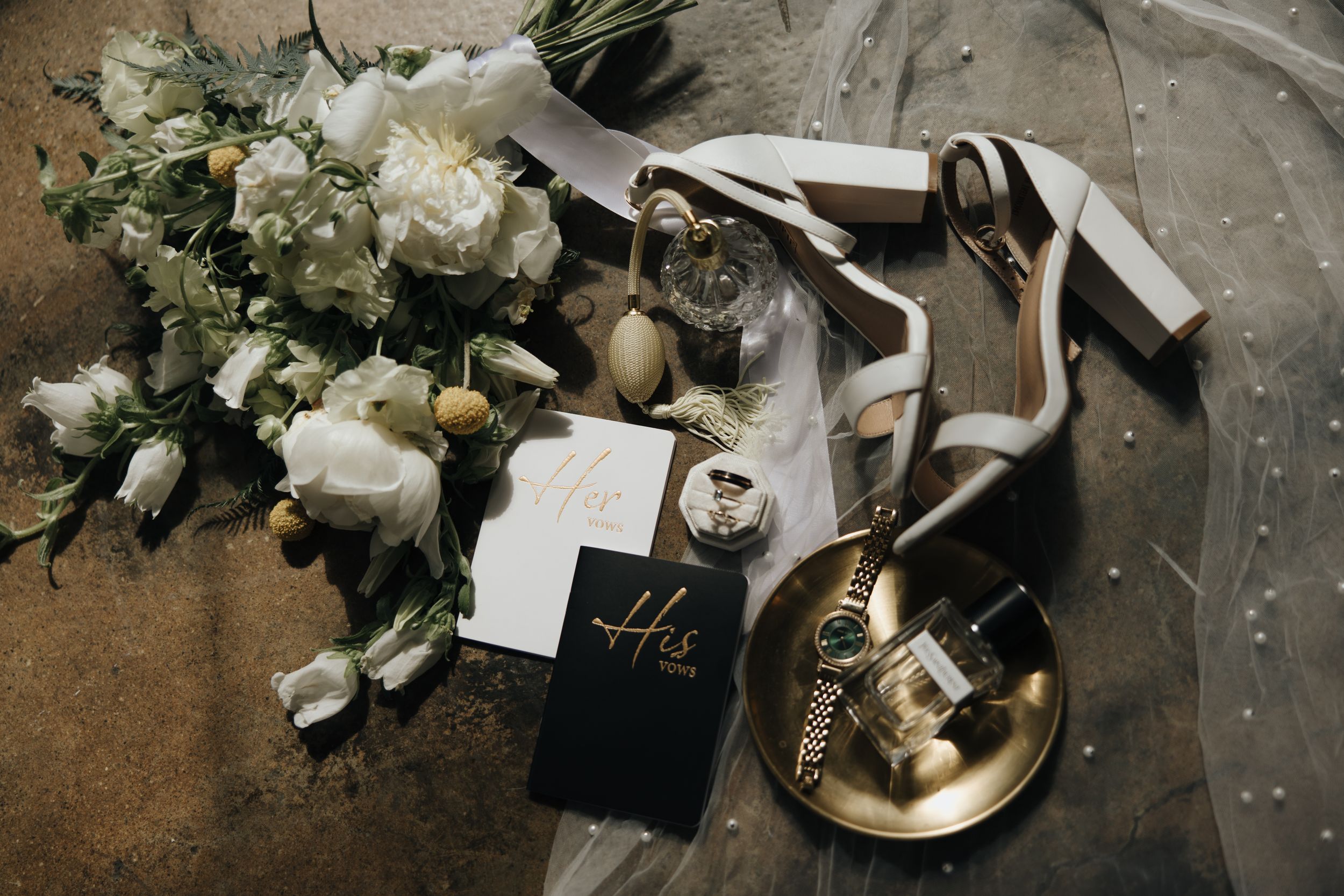 Wedding details flat lay with white floral bouquet, high heels, and invitation on concrete surface.