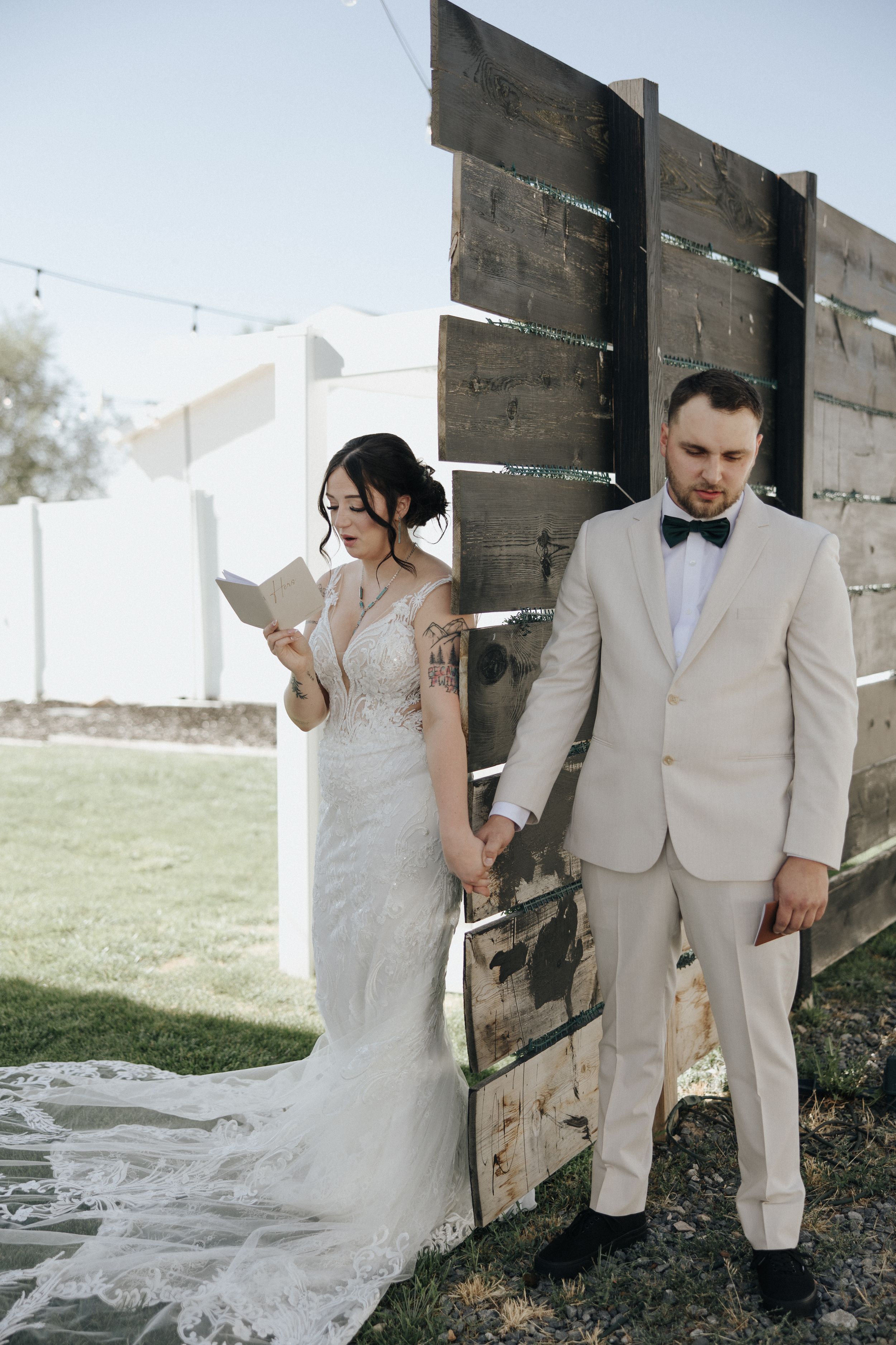 A romantic wedding couple poses together in front of a rustic barn wall with string lights overhead.