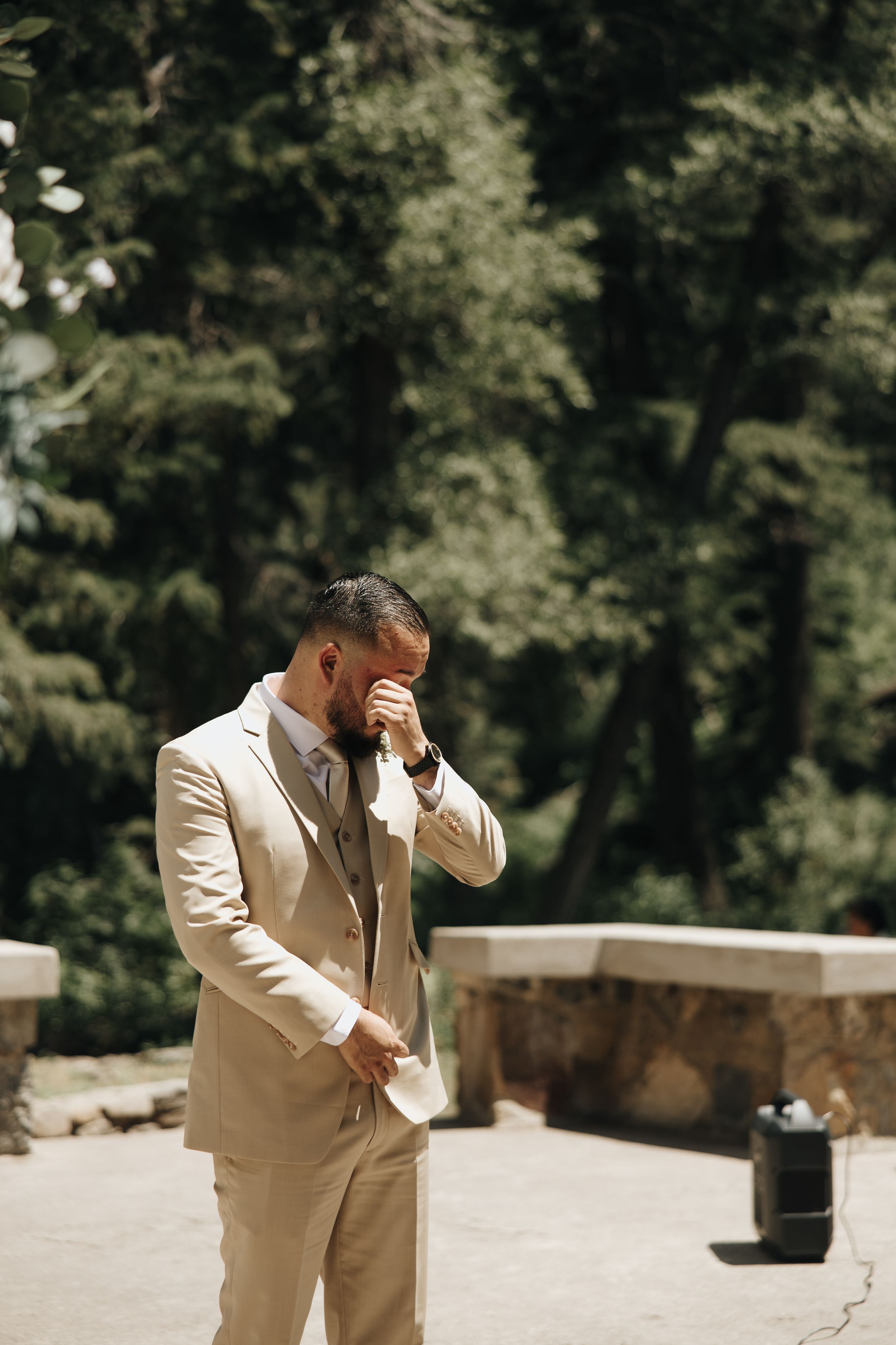 Groom in beige suit wipes tears during emotional first look moment in outdoor garden setting.