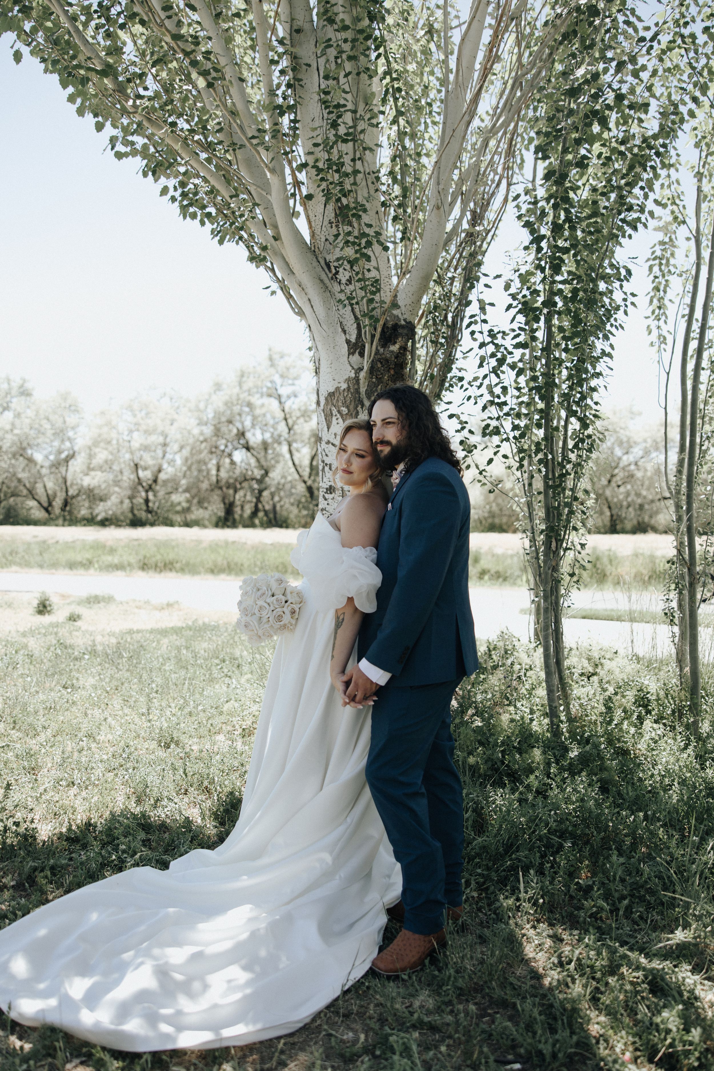 A couple shares an intimate moment under a towering white birch tree on their wedding day in a rustic outdoor setting.