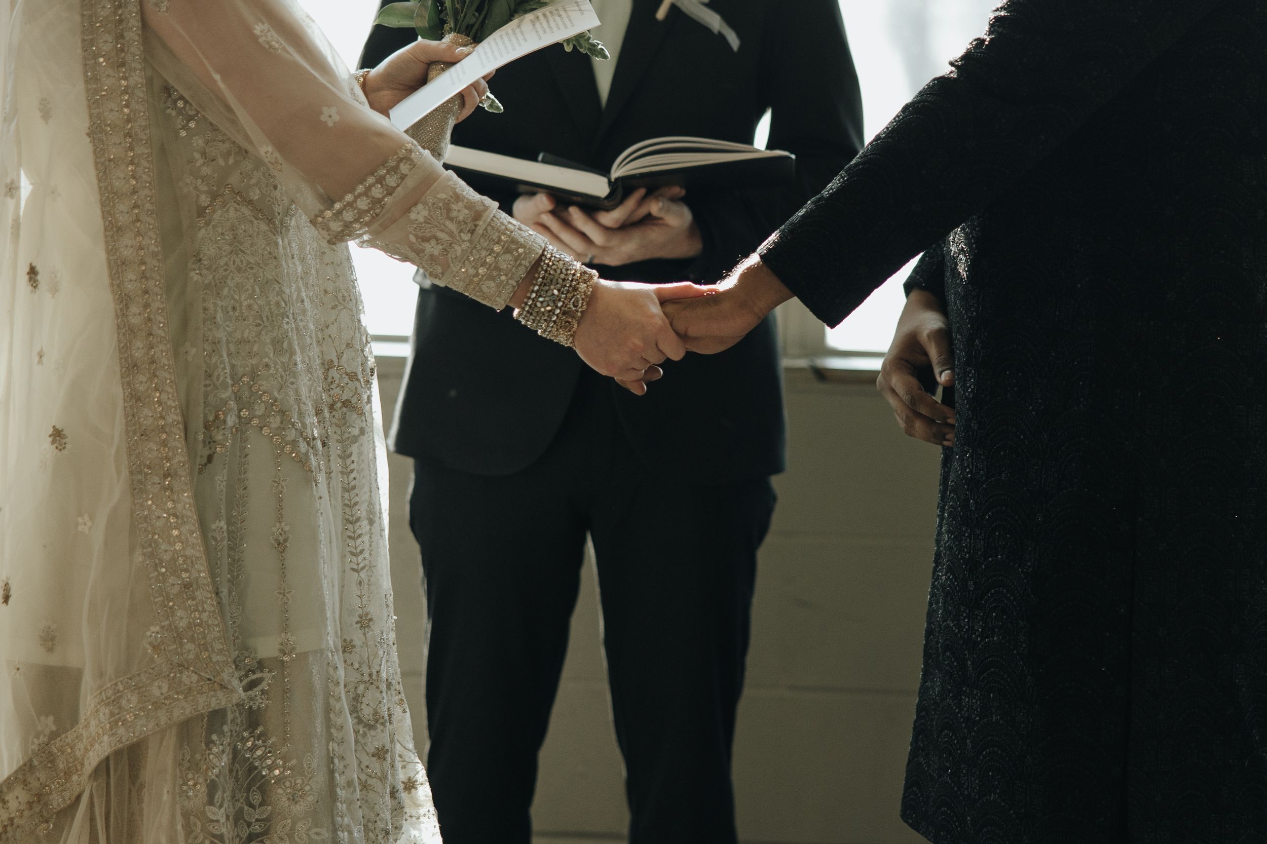 A couple holding hands during a wedding ceremony, with the bride in a lace dress and groom in a black suit.
