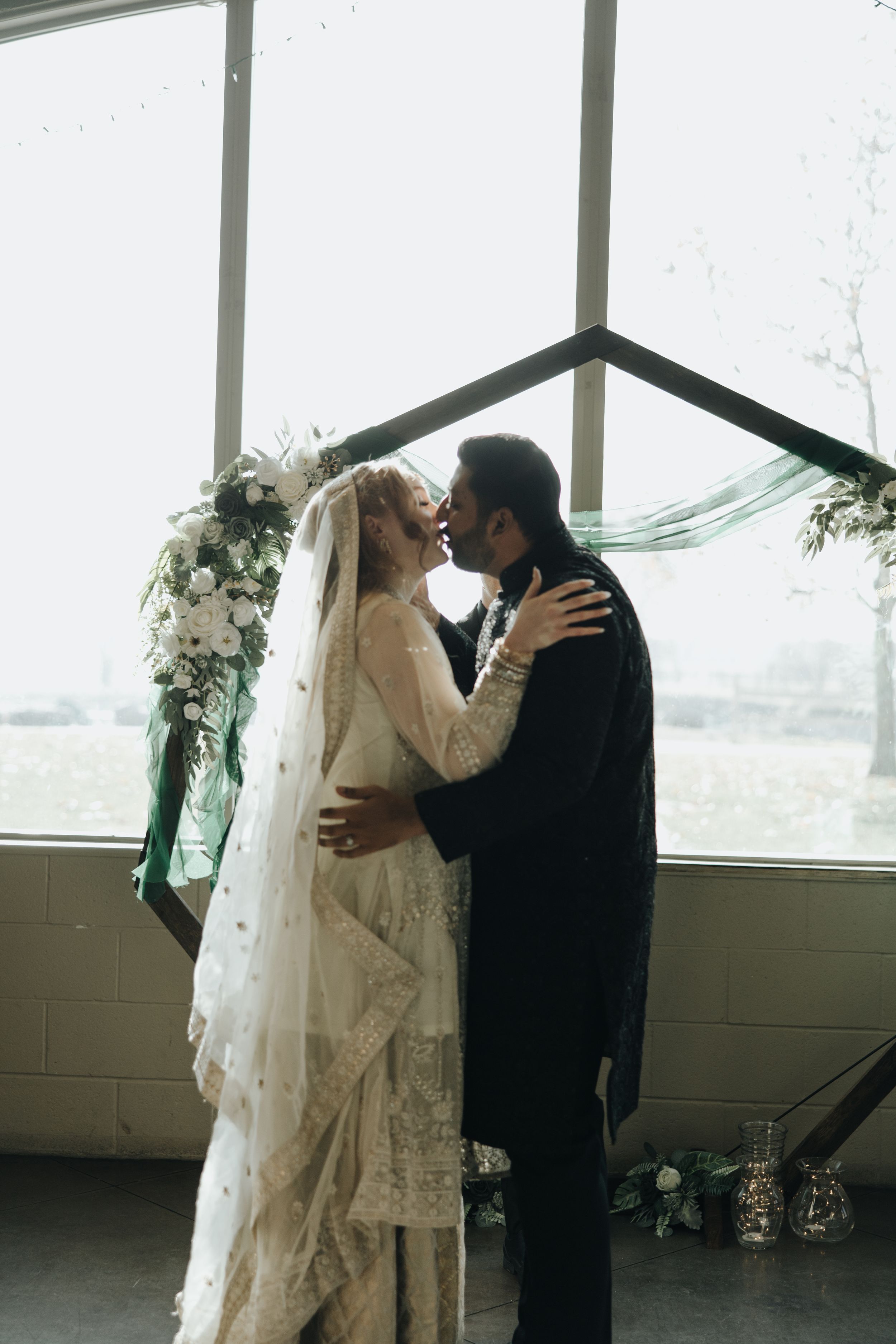 A romantic wedding embrace captured near large windows beneath a floral arch with a long flowing veil.