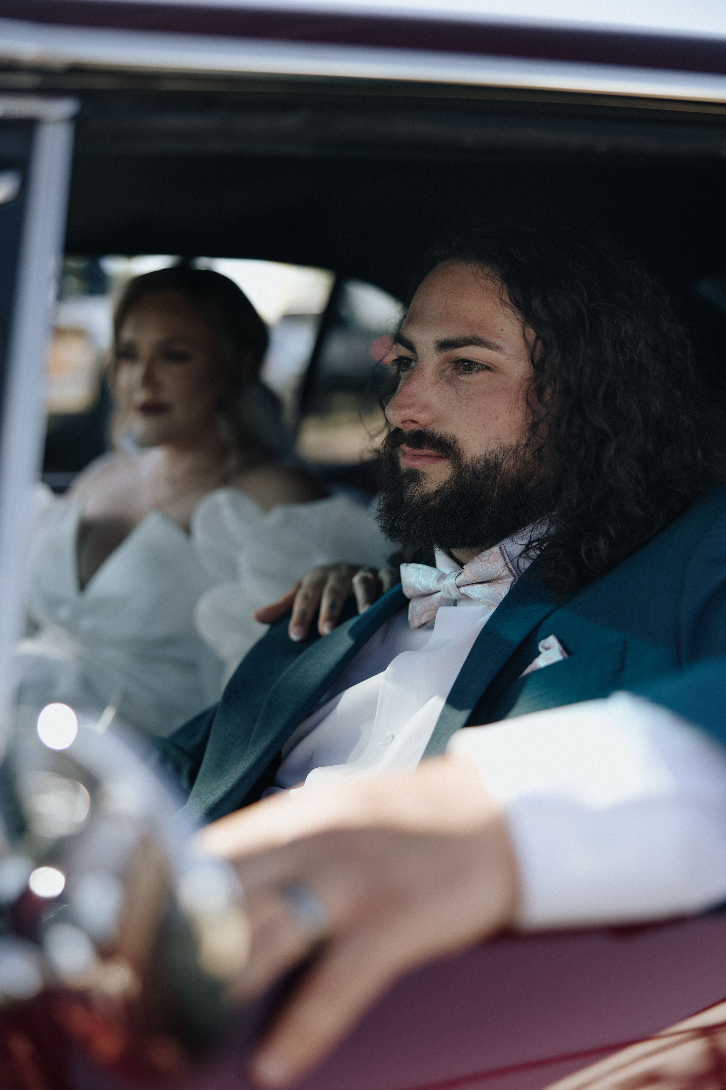 A couple dressed in formal wedding attire sit together in a car, with the passenger in white and driver wearing a bow tie.